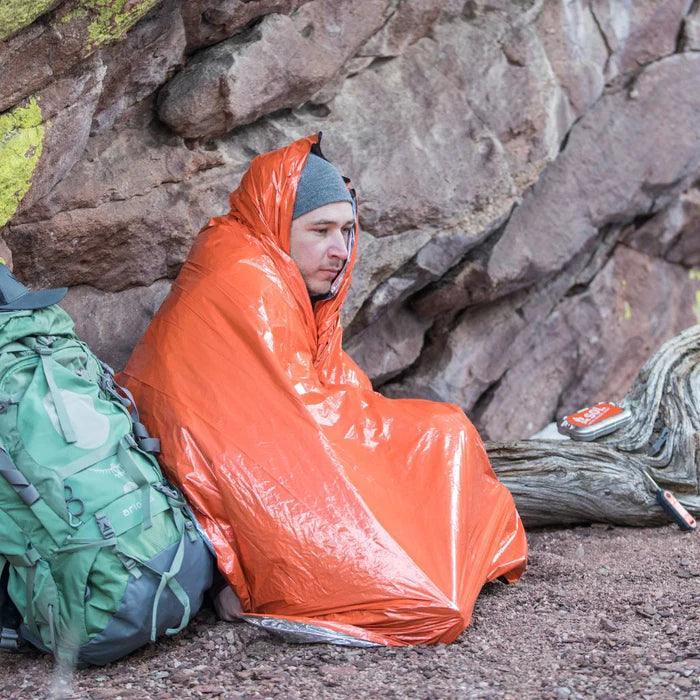Man wrapped in orange emergency blanket by rock wall with hiking backpack outdoors