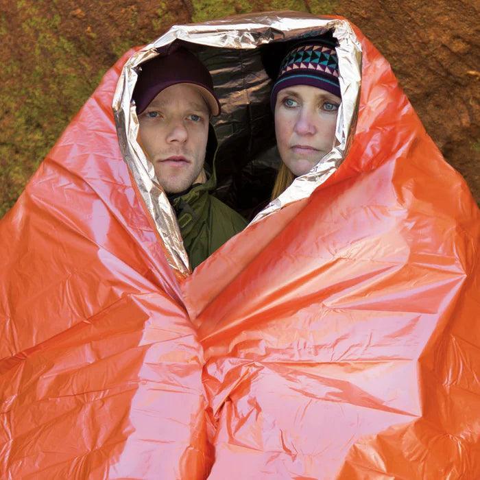 Two people sheltered in an orange emergency blanket outdoors near rocks