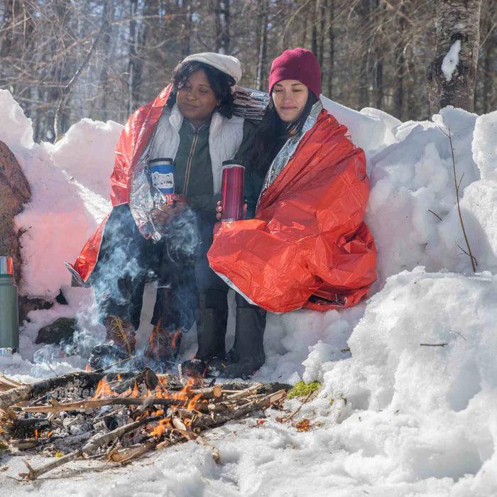 Two people sit by a campfire in snow, wrapped in orange SOL emergency blankets outdoors.