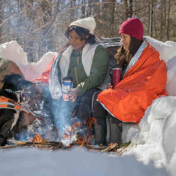 Two women with emergency blankets sit by a winter campfire with a dog in snowy woods