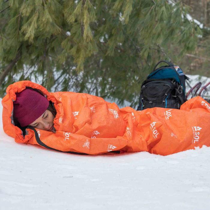 Camper in orange SOL Escape Bivvy sleeping bag on snow with backpack, winter outdoors
