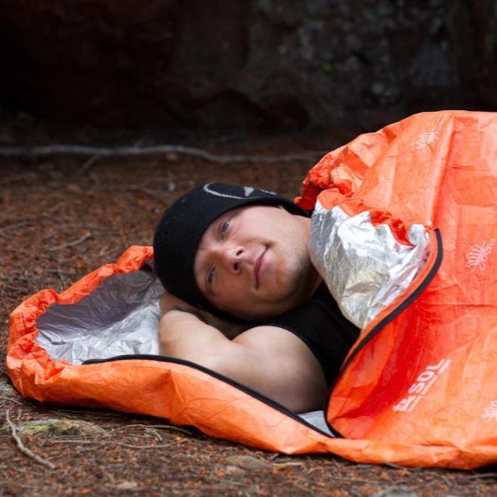 Man in orange SOL Escape Bivvy sleeping bag outdoors on forest floor