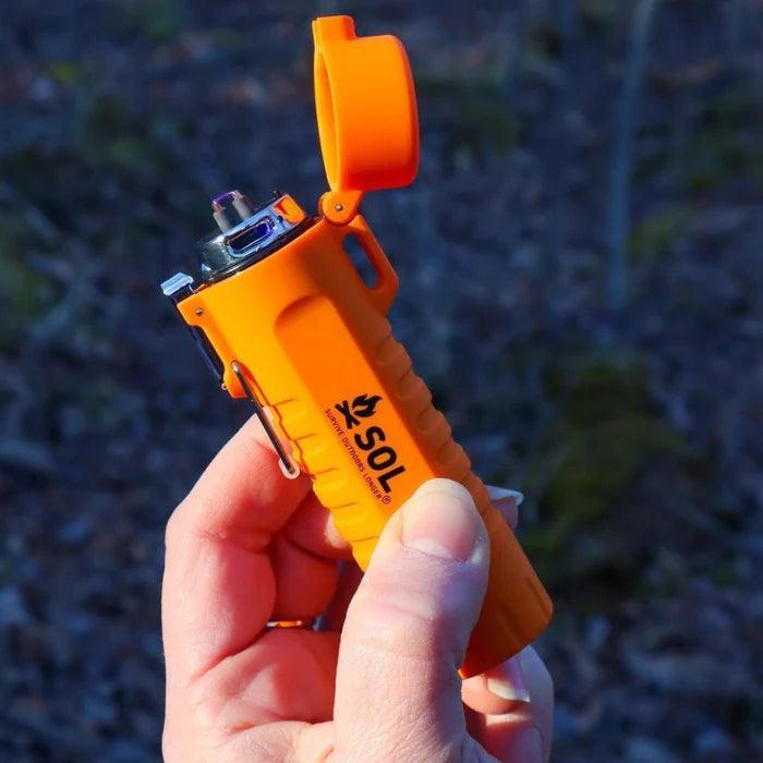 Hand holding orange SOL Fire Lite Fuel-Free Lighter outdoors, survival gear focus.