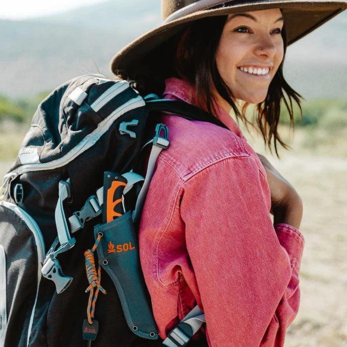Woman outdoors with backpack carrying SOL Stoke Pivot Knife and Saw, pink jacket, wide-brim hat