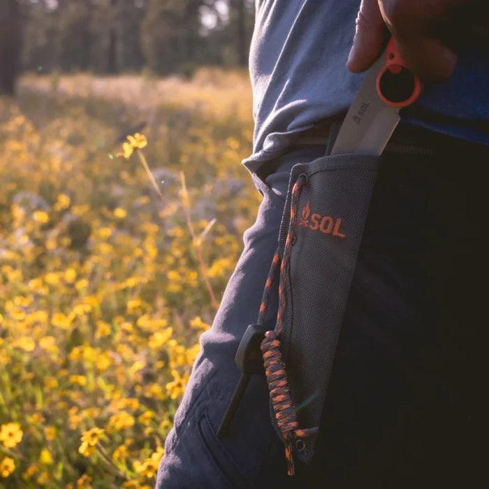 SOL Pivot Knife in sheath on belt outdoors with wildflowers and person in blue shirt