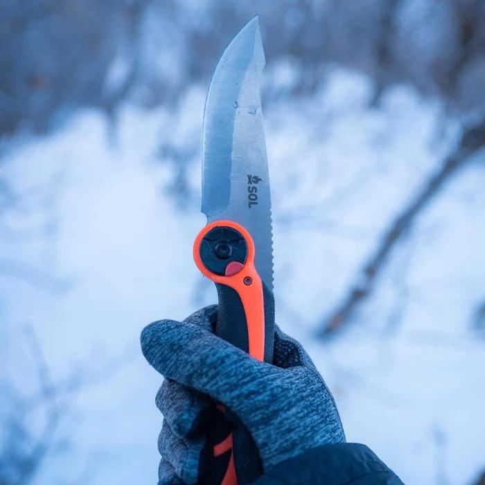 Hand holding SOL folding knife with orange and black handle in snowy outdoor setting