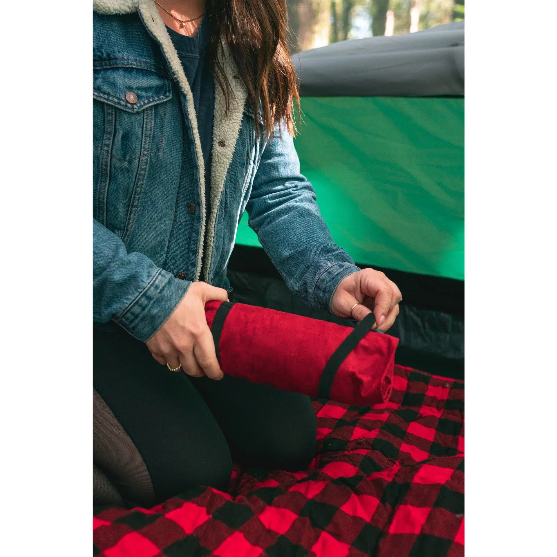 Person in denim jacket rolling a red plaid camping blanket inside a tent