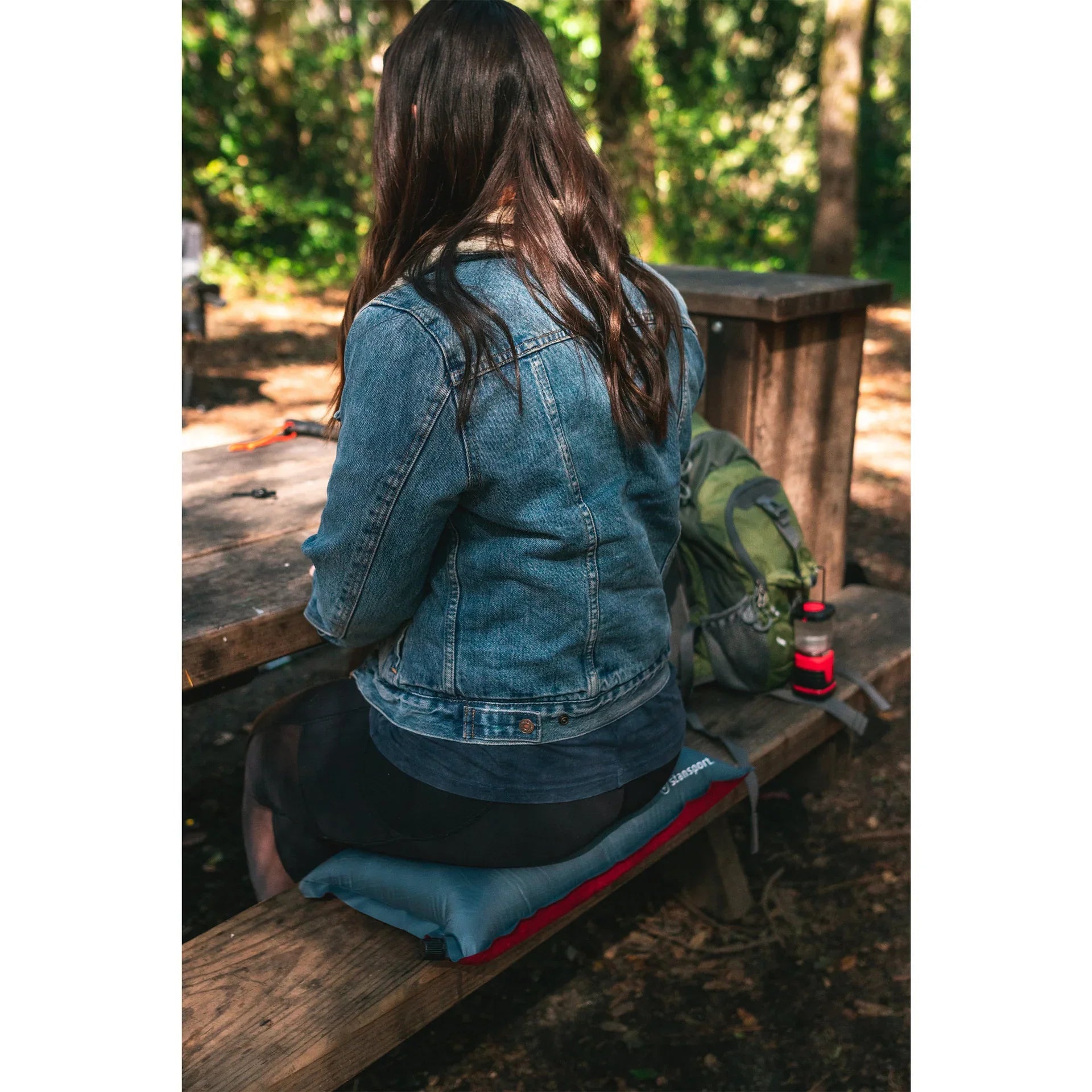 Woman in denim jacket sits on portable seat pad at picnic table in forest campsite