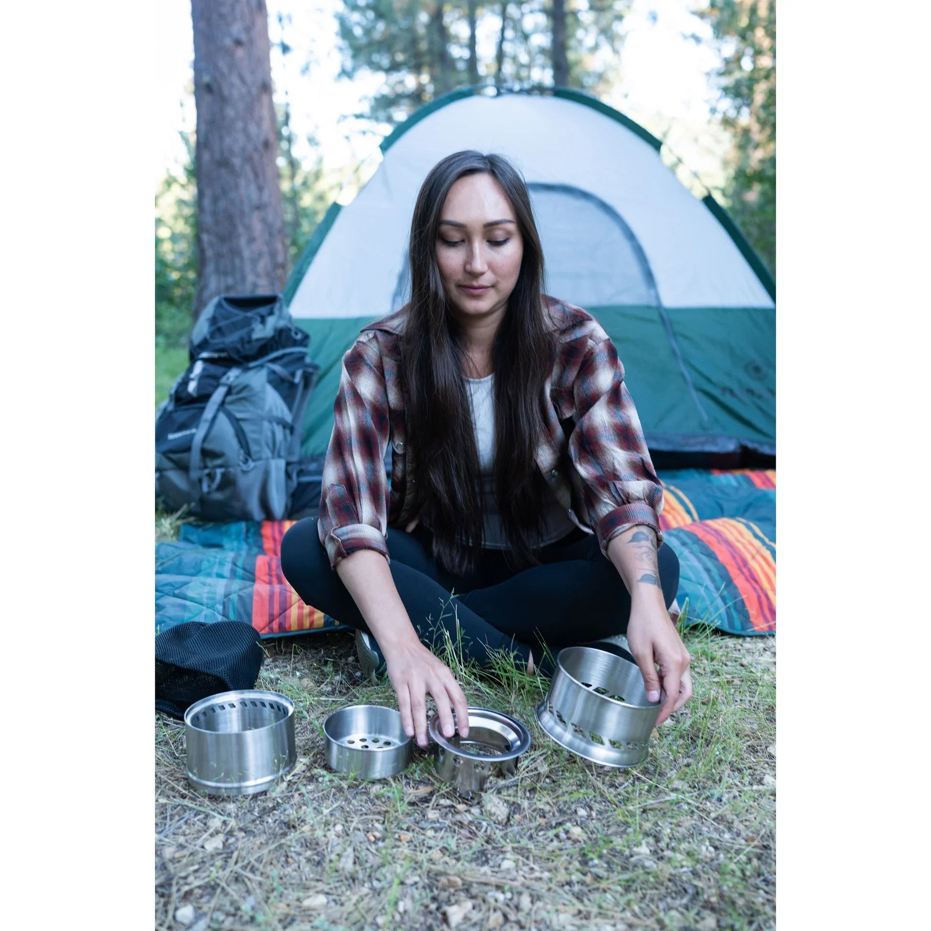 Woman assembling camping stove outdoors near tent and backpack in forest setting