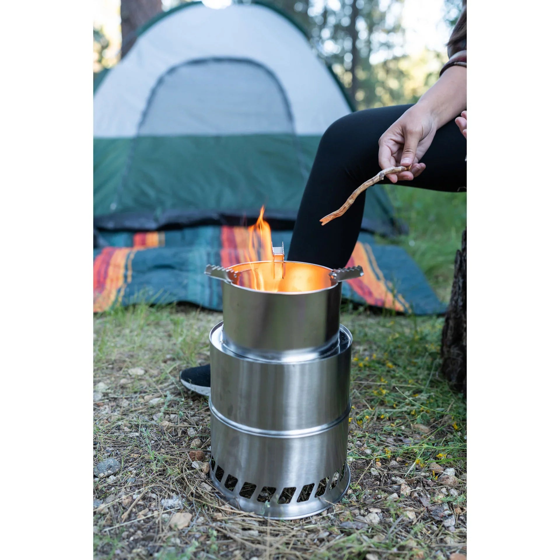 Camping stove burning outside green tent, person adding stick to fire, outdoor camping gear