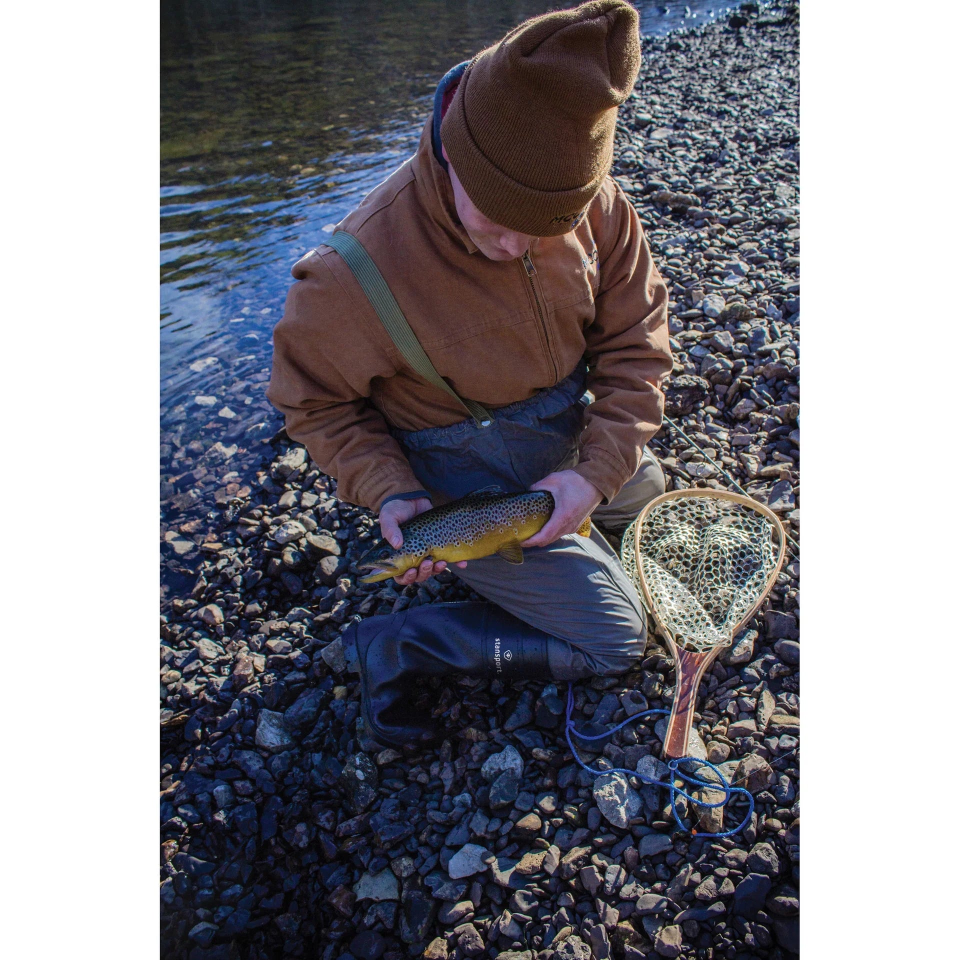 Fisherman kneeling on rocky riverbank holding trout with fishing net and waders