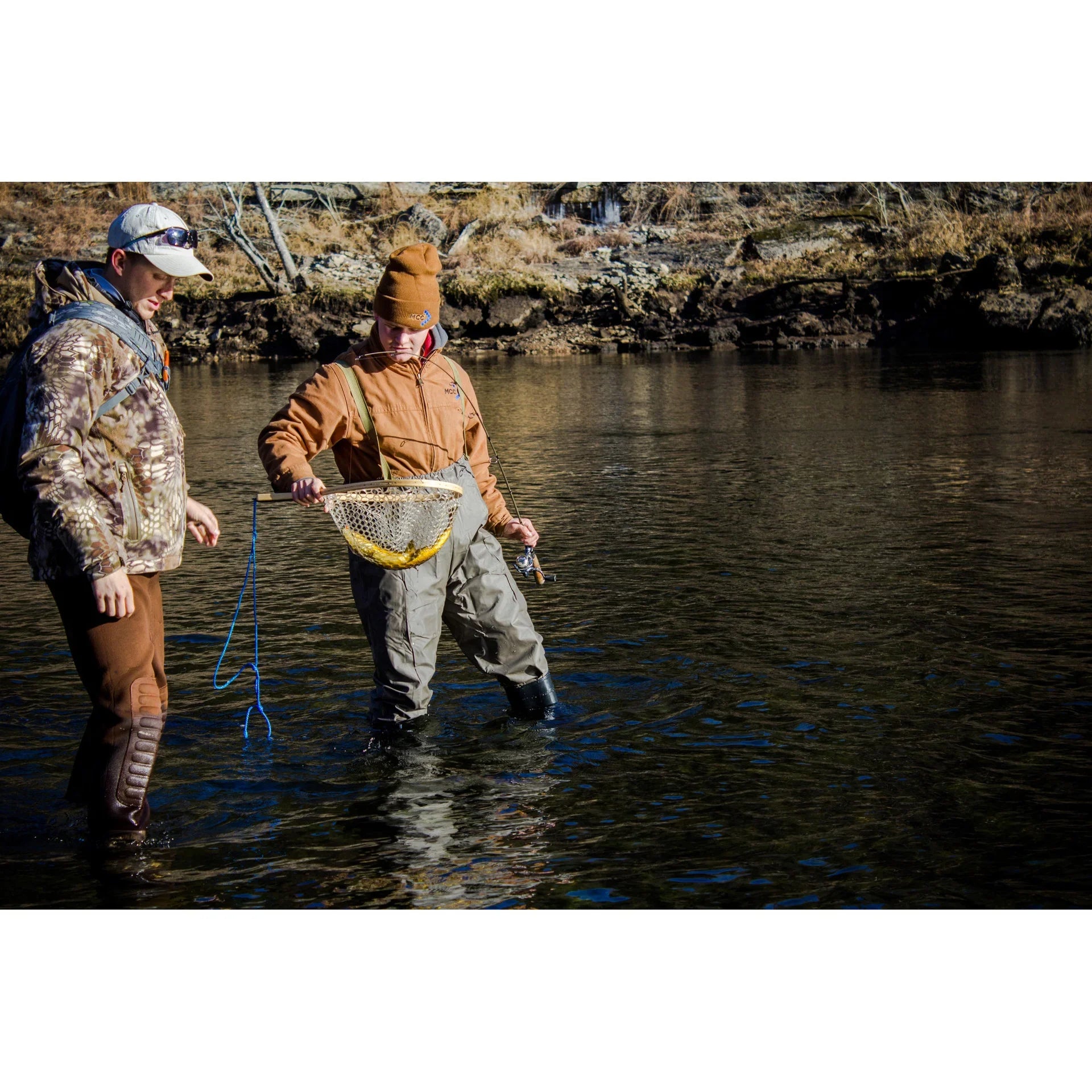Two men fly fishing in river, wearing waders and outdoor gear, holding a fishing net