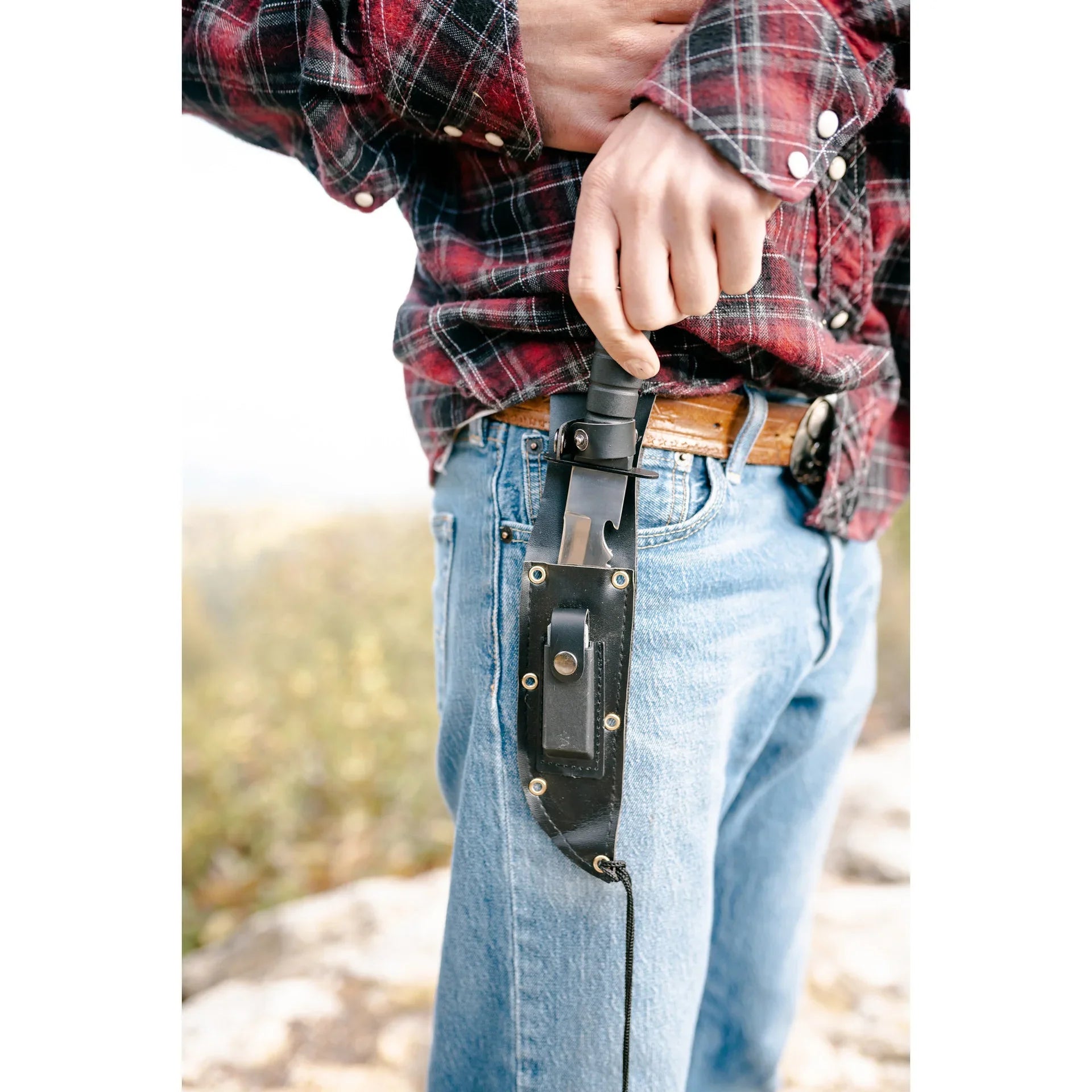 Person in plaid shirt and jeans drawing survival knife from black sheath on belt outdoors