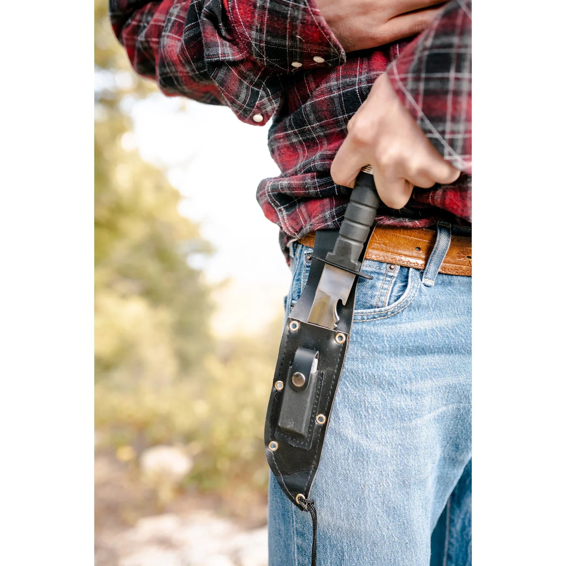Person drawing survival knife from black sheath, wearing plaid shirt and jeans outdoors