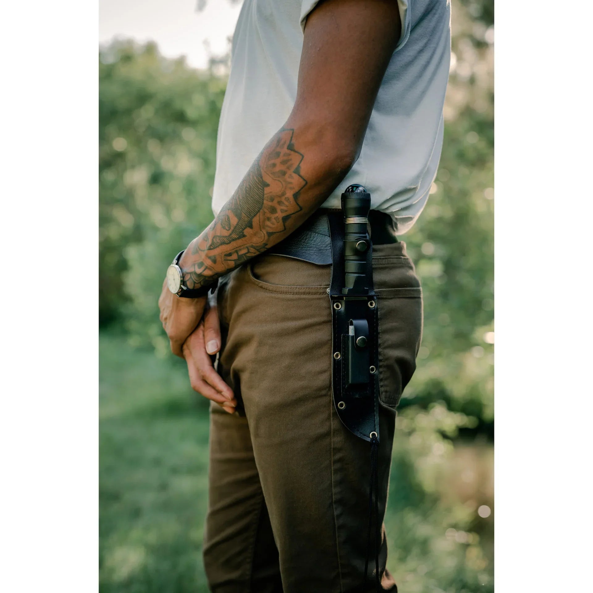 Tattooed arm, tan pants, sheath knife on belt, outdoor setting, survival gear.