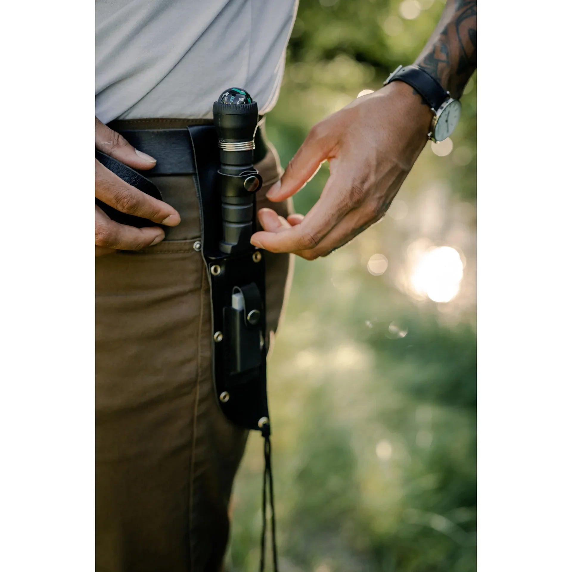 Man wearing tactical knife and flashlight pouch on belt outdoors.