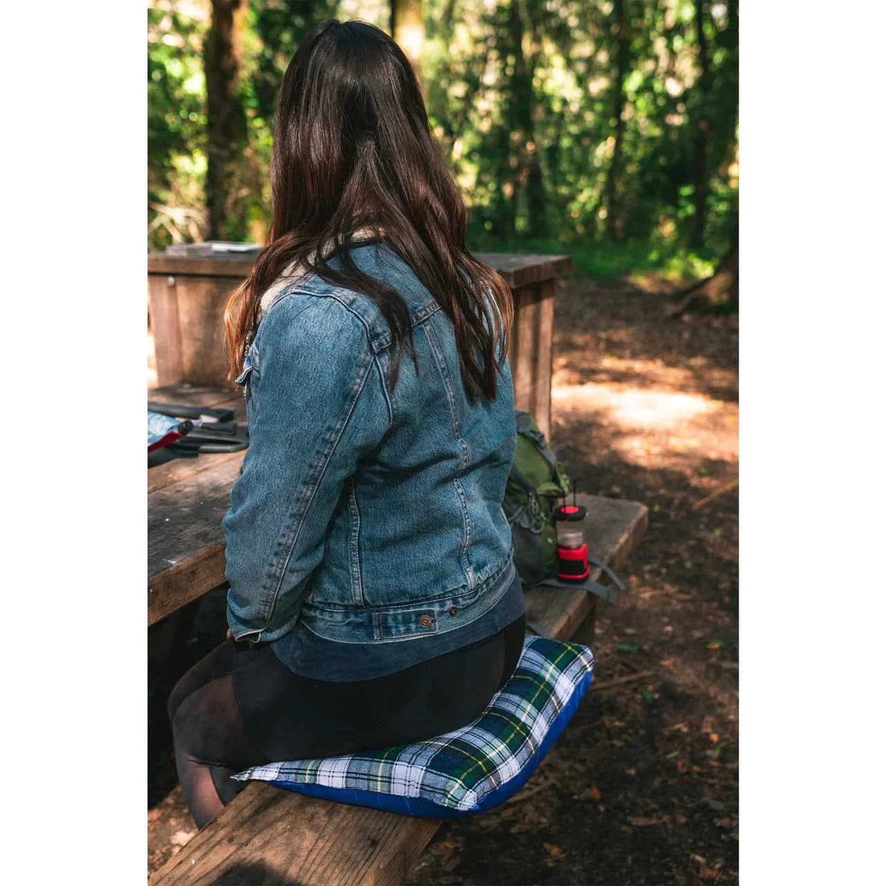 Woman in denim jacket sitting on plaid Stansport washable camp pillow at a wooden picnic table outdoors