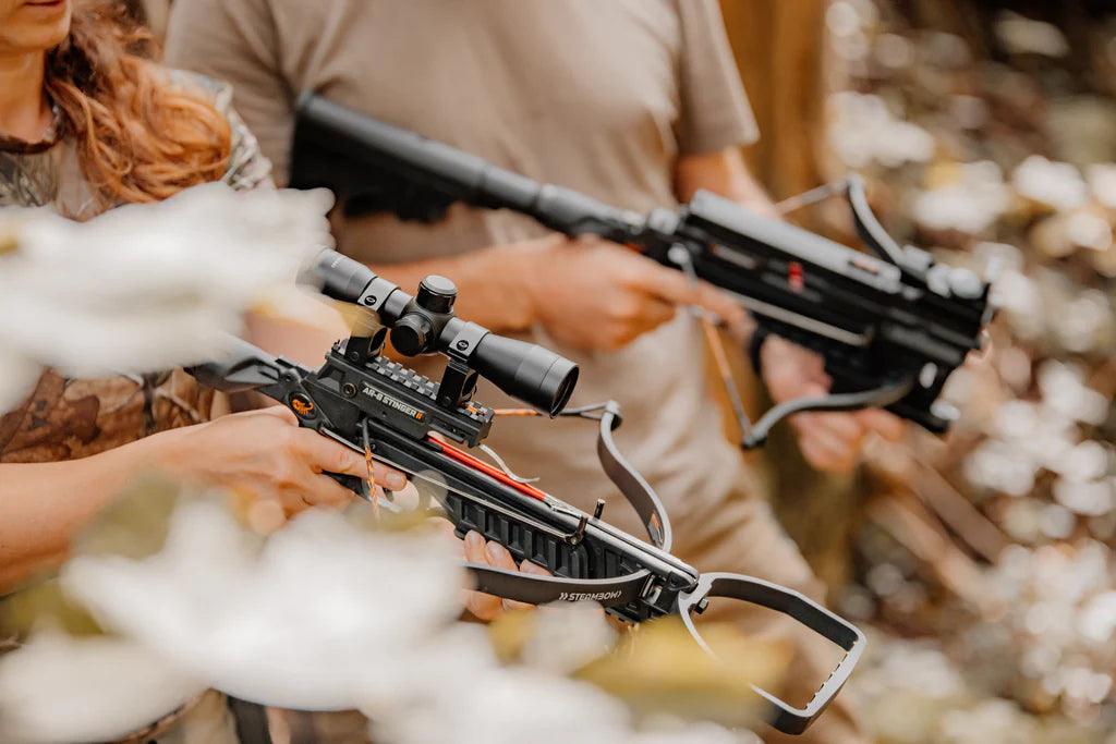 Two people outdoors holding black crossbows with scopes in a forest setting