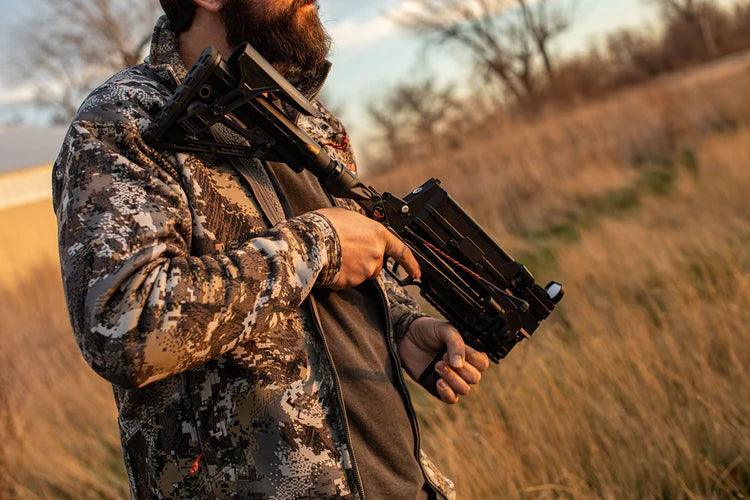 Man in camo jacket holding folding stock crossbow outdoors in tall grass at sunset