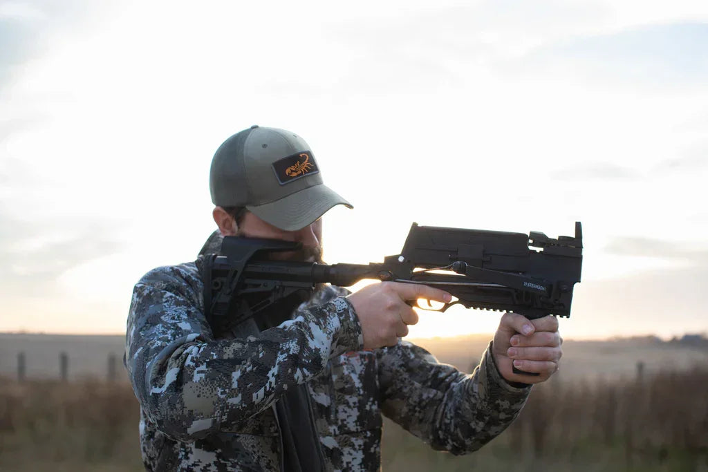 Man in camo jacket aiming black Stinger crossbow outdoors at sunset