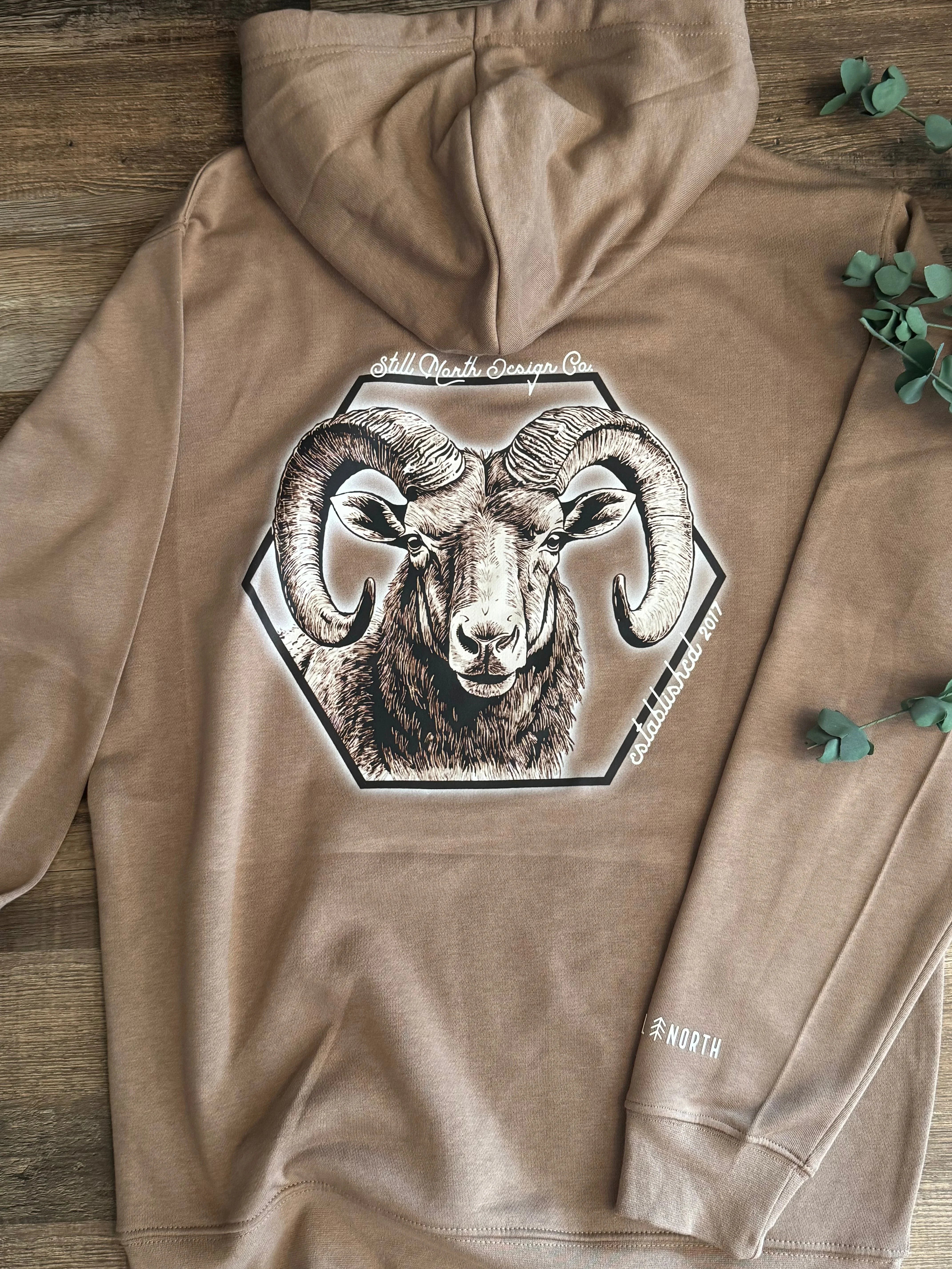 Brown hoodie with ram graphic and Still North Design Co. logo on wooden surface
