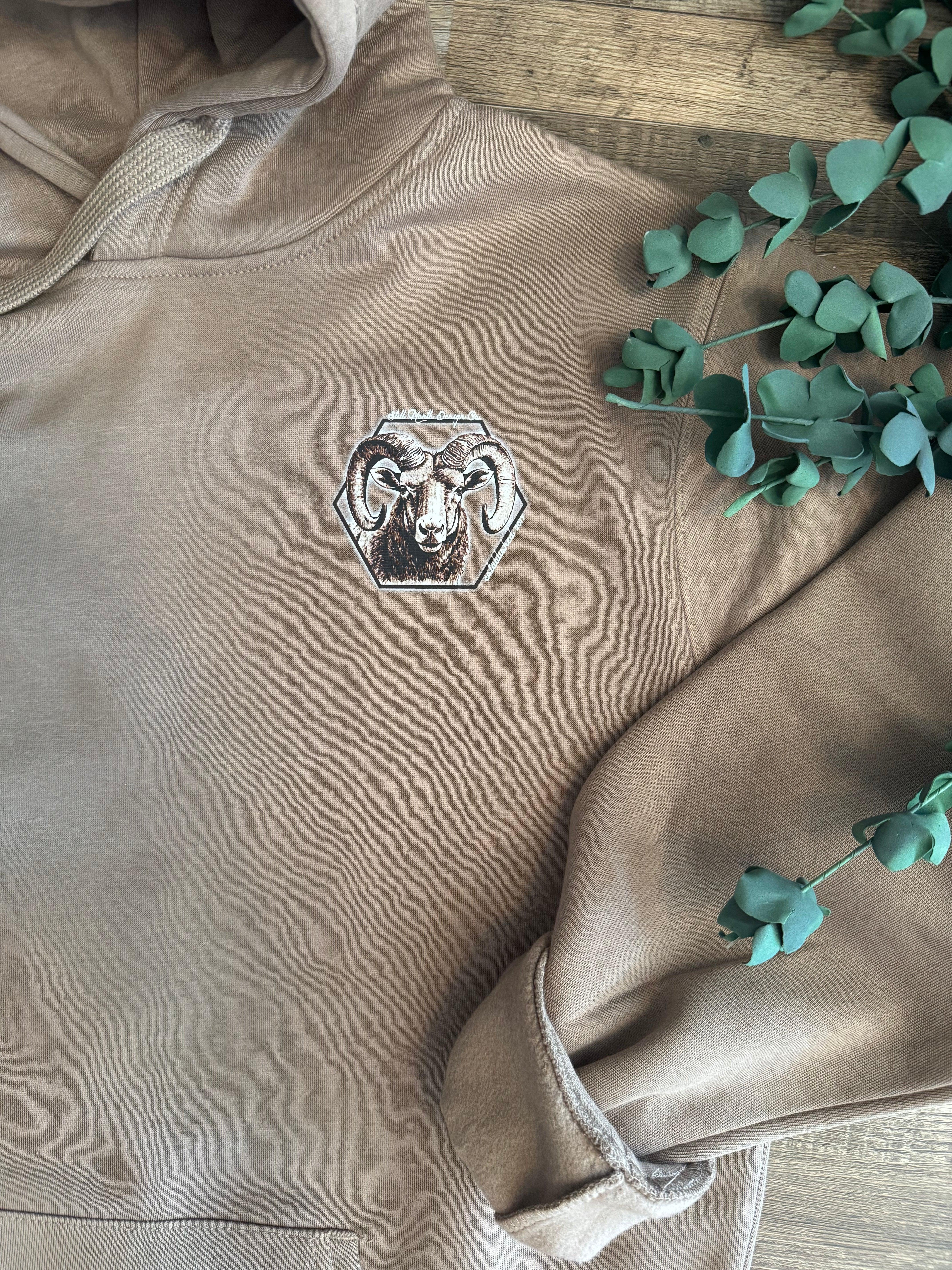 Brown hoodie with ram head graphic, wooden background, green eucalyptus leaves