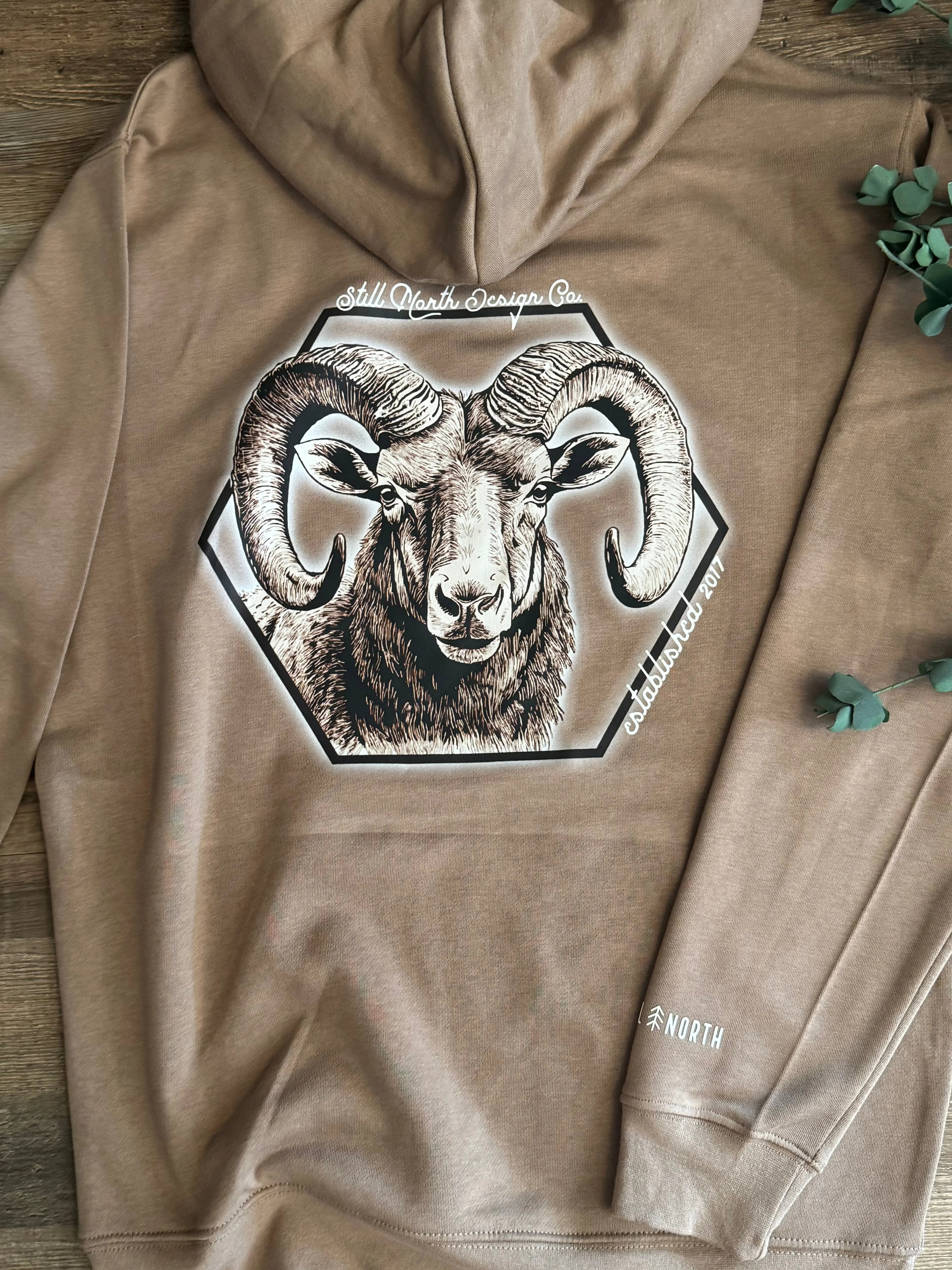 Brown hoodie with ram graphic and Still North Design Co. logo on wooden surface