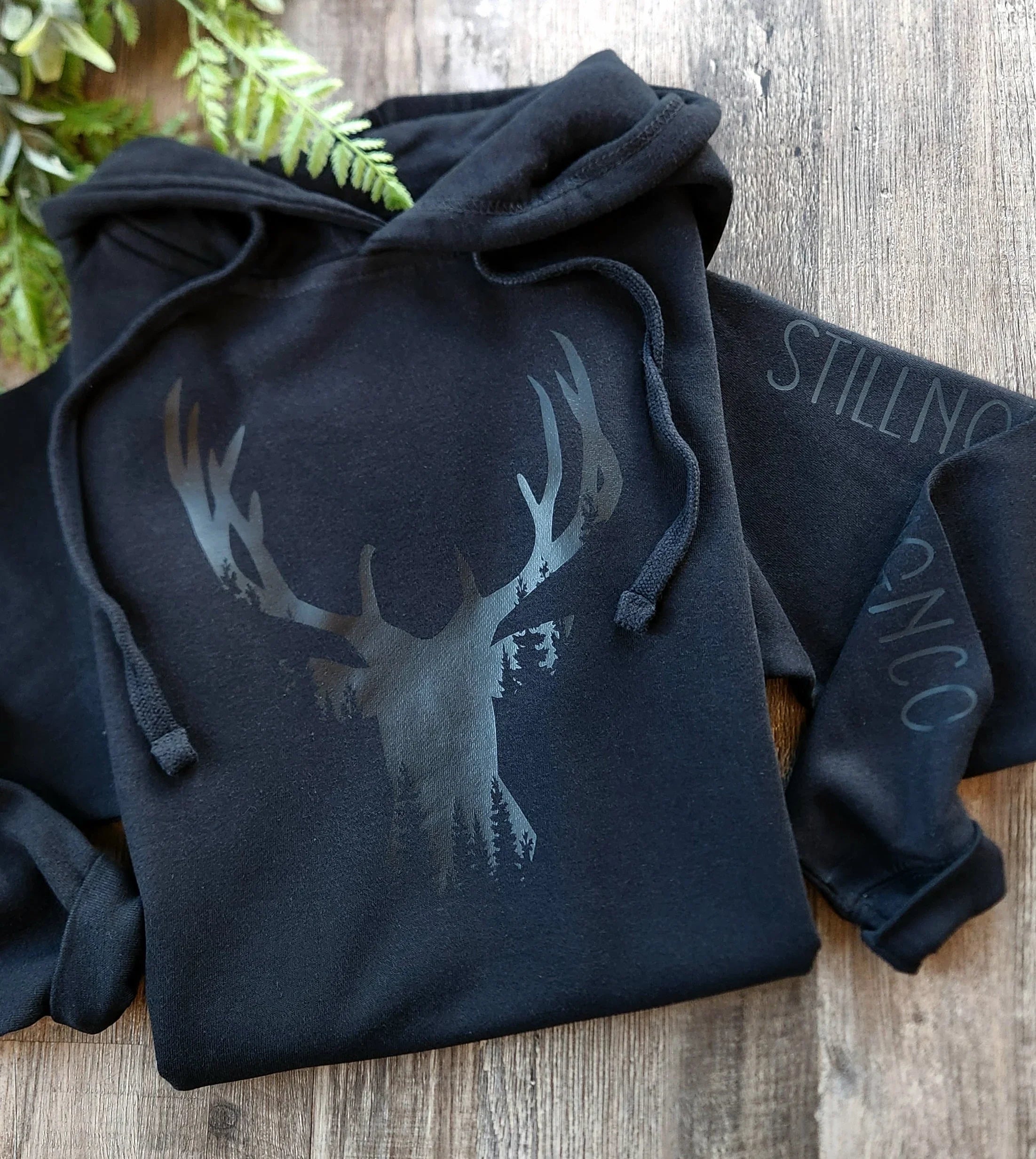 Black hoodie with glossy deer and forest design on wooden surface, minimalist nature apparel
