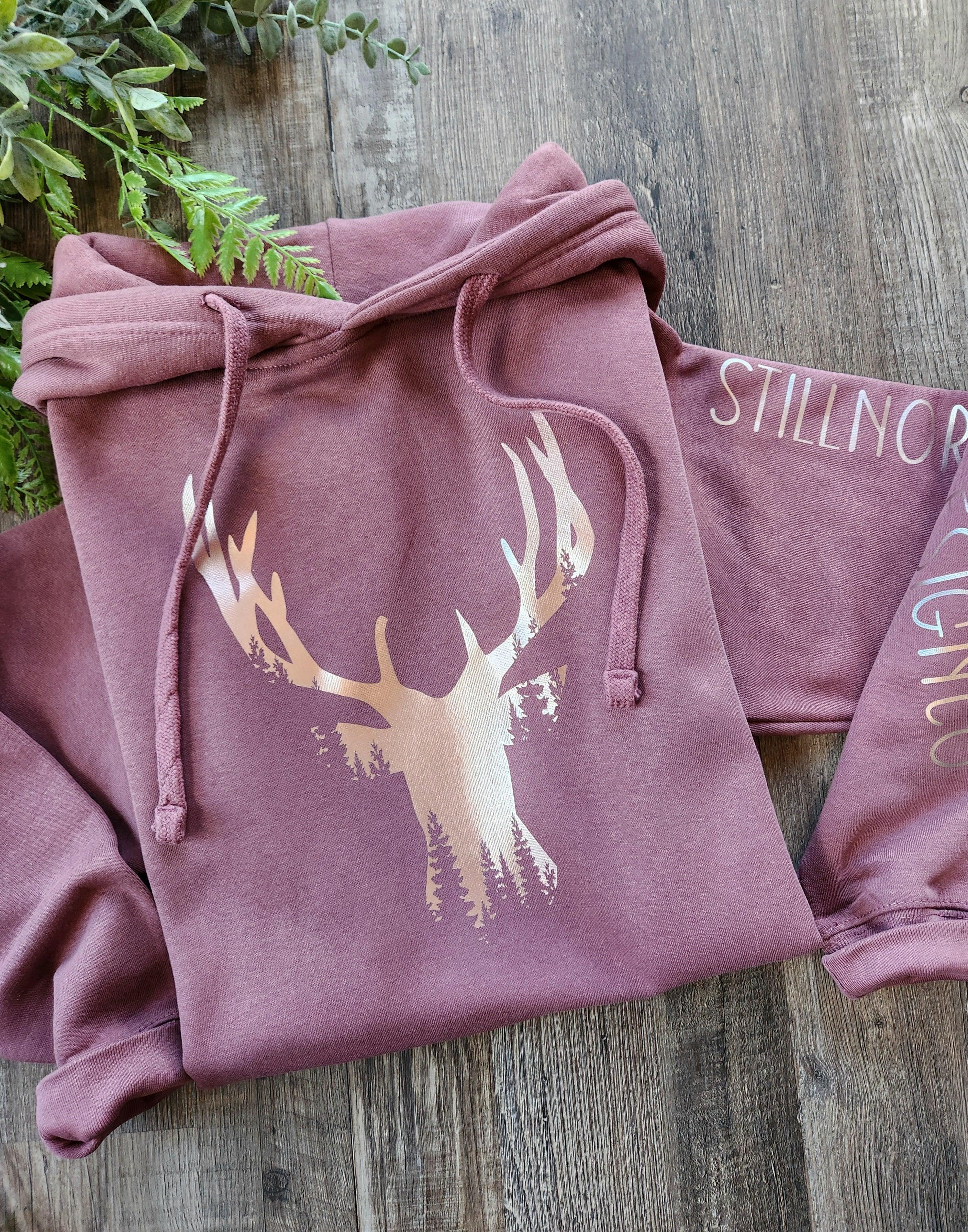 Mauve hoodie with stag and forest graphic, STILLNOR text on sleeve, on wooden surface
