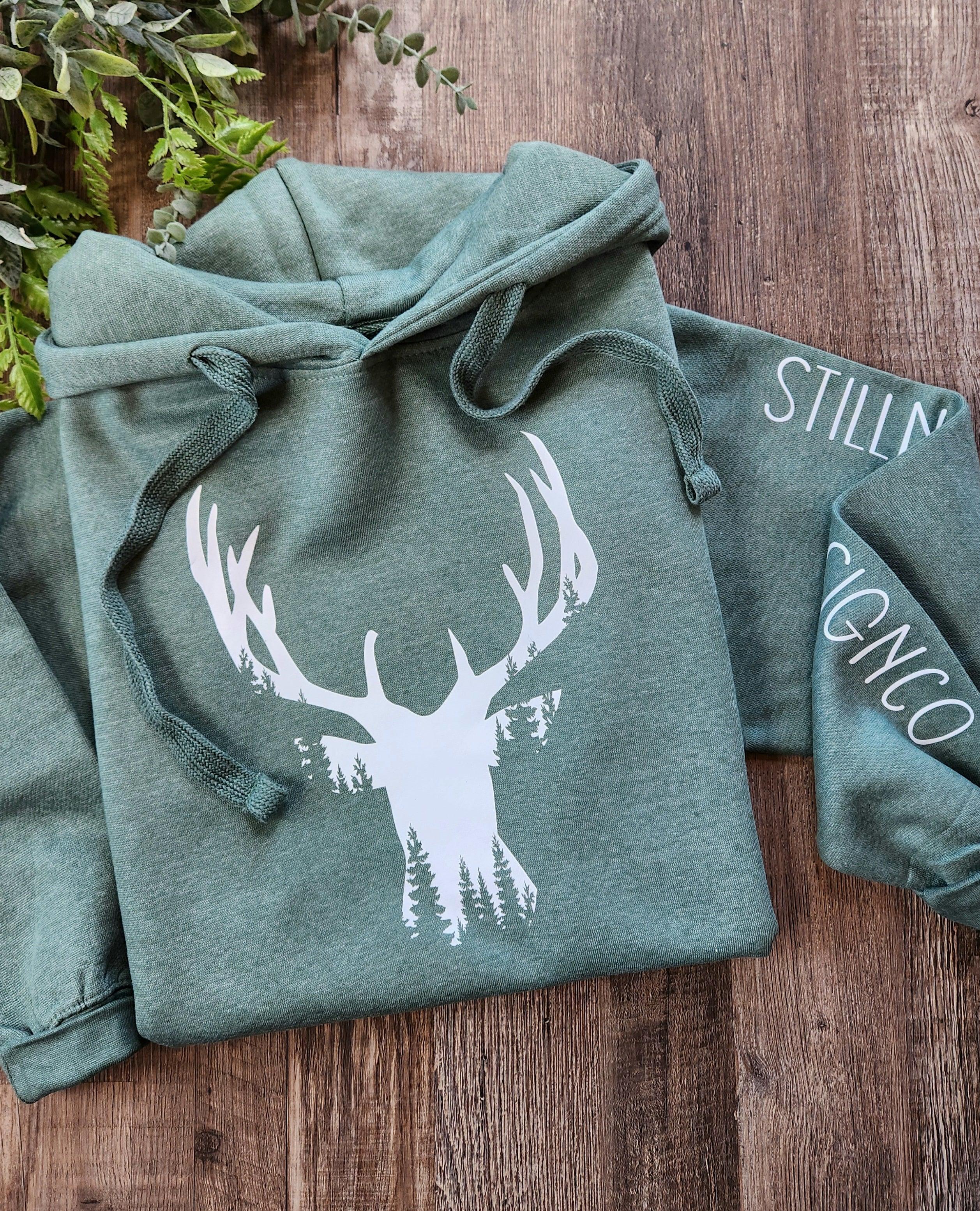 Green hoodie with white deer head and forest print on wood background, rustic apparel