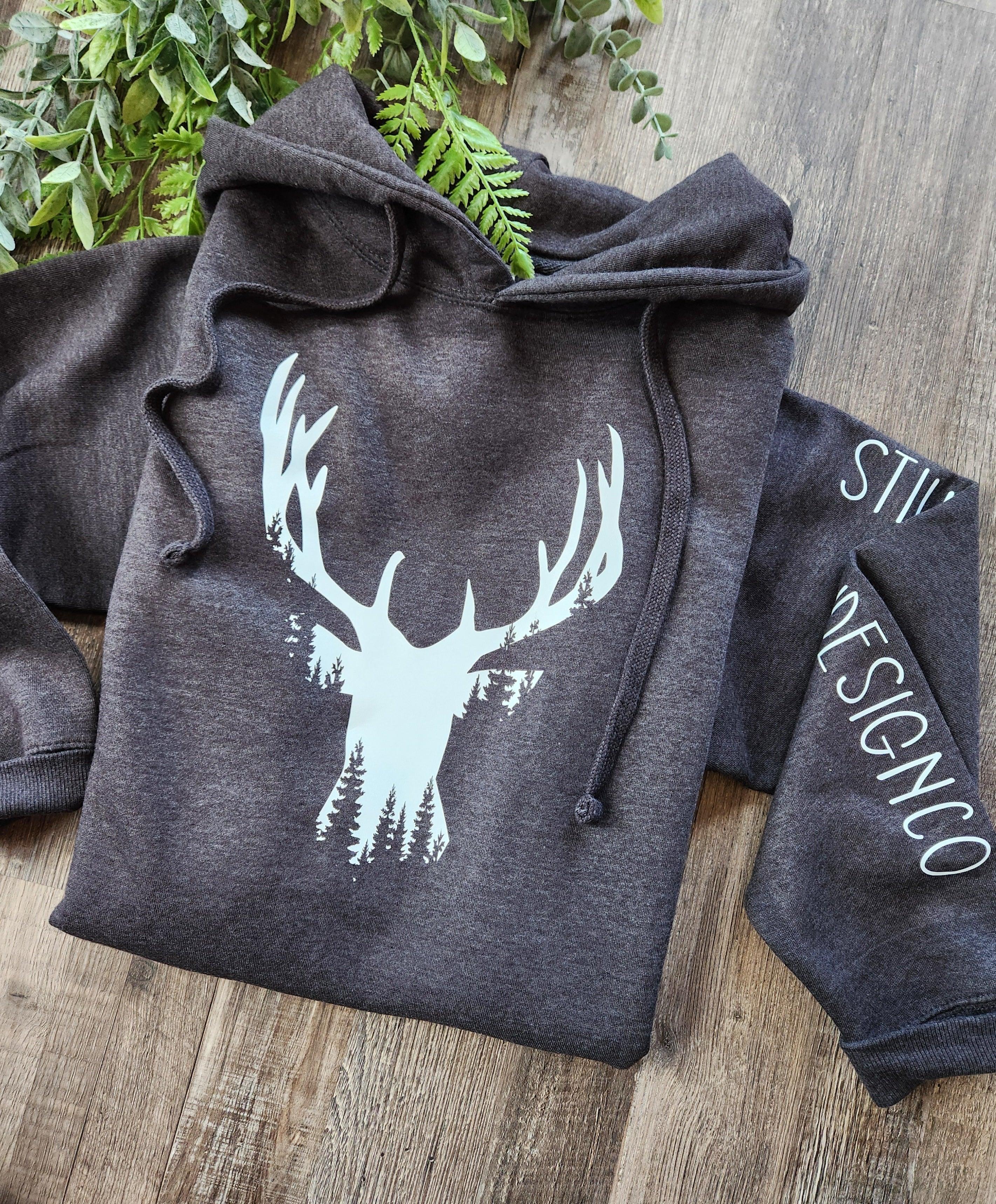 Dark grey hoodie with white deer antler and tree design, laid on wood surface with greenery.