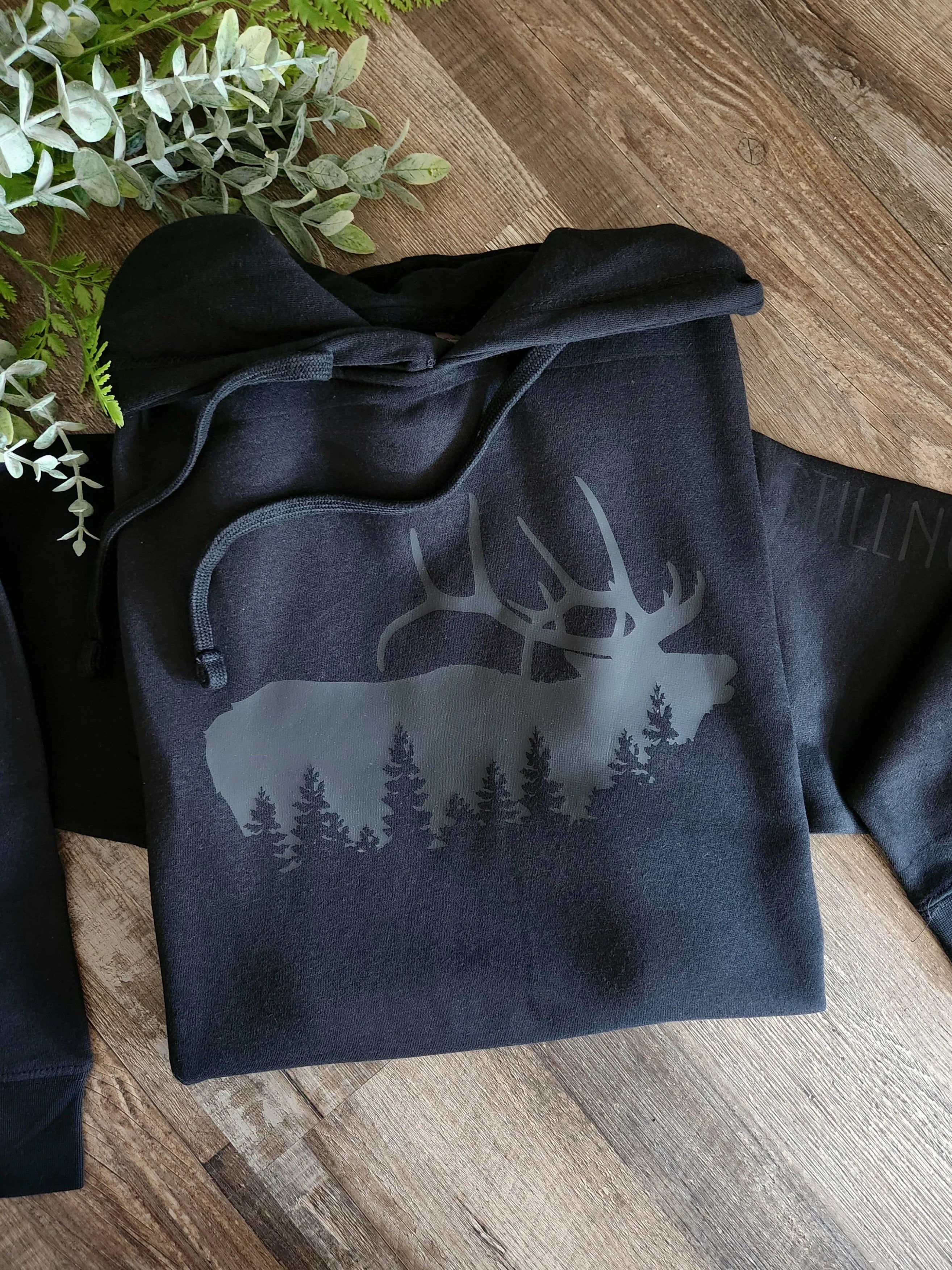 Black hoodie with elk and forest silhouette graphic on wooden surface with green leaves