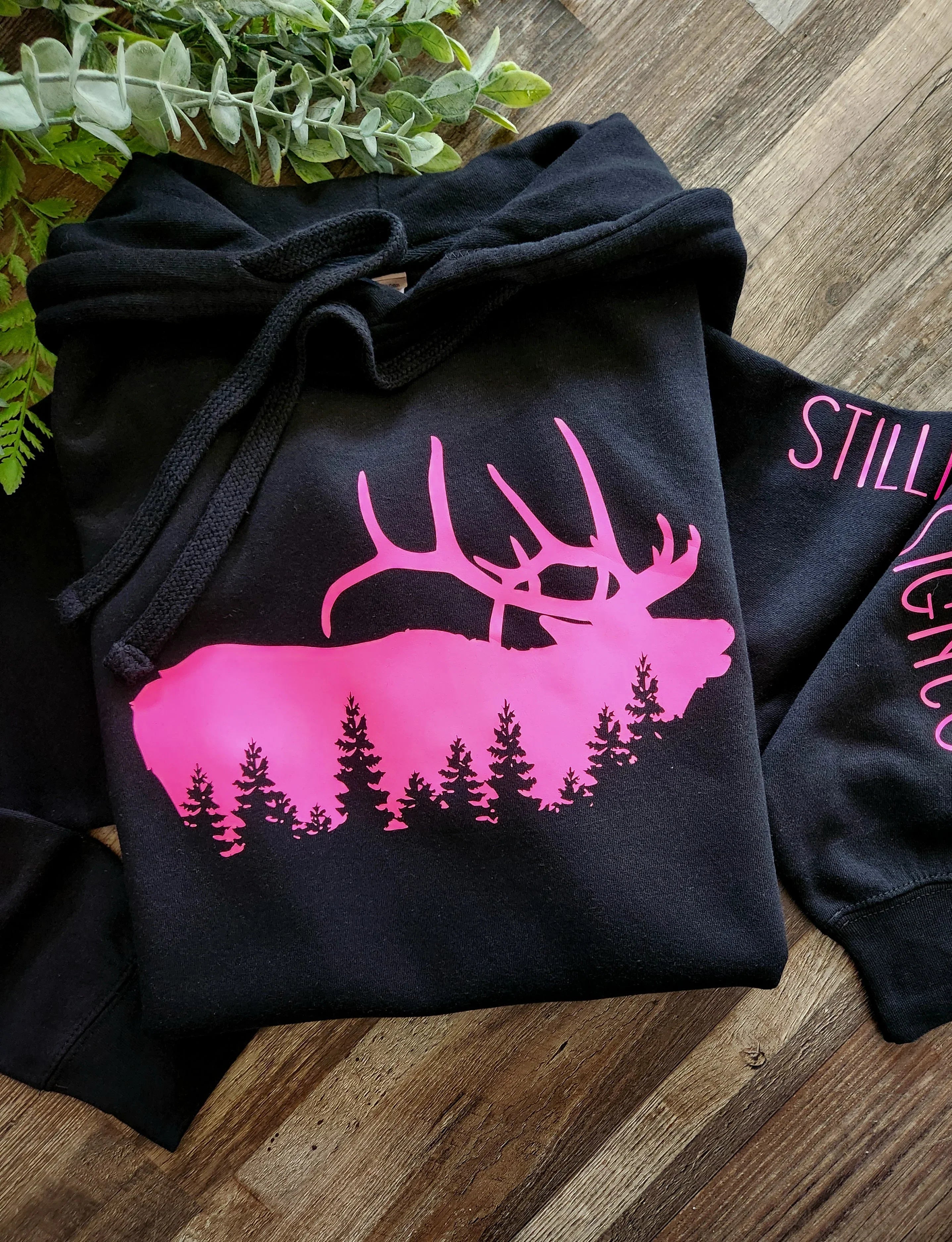 Black hoodie with pink elk and pine tree graphic, rustic wood background, greenery decor