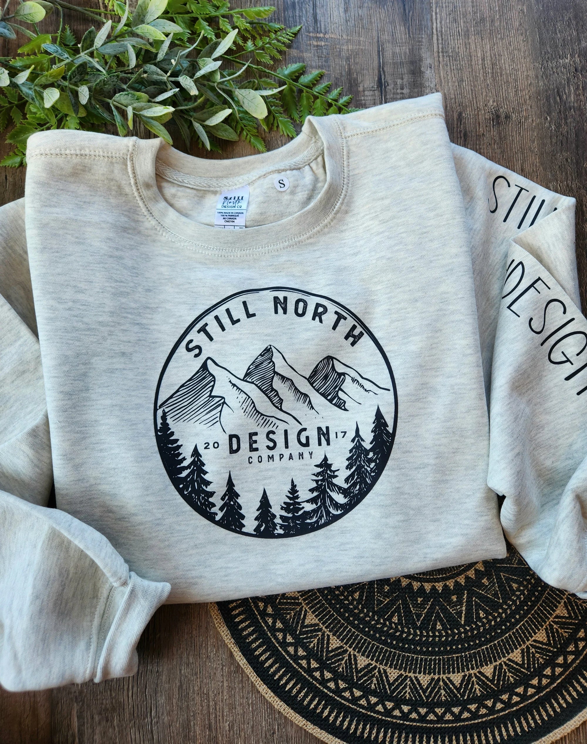 Beige crewneck sweatshirt with Still North Design Company mountain and trees graphic on wood table