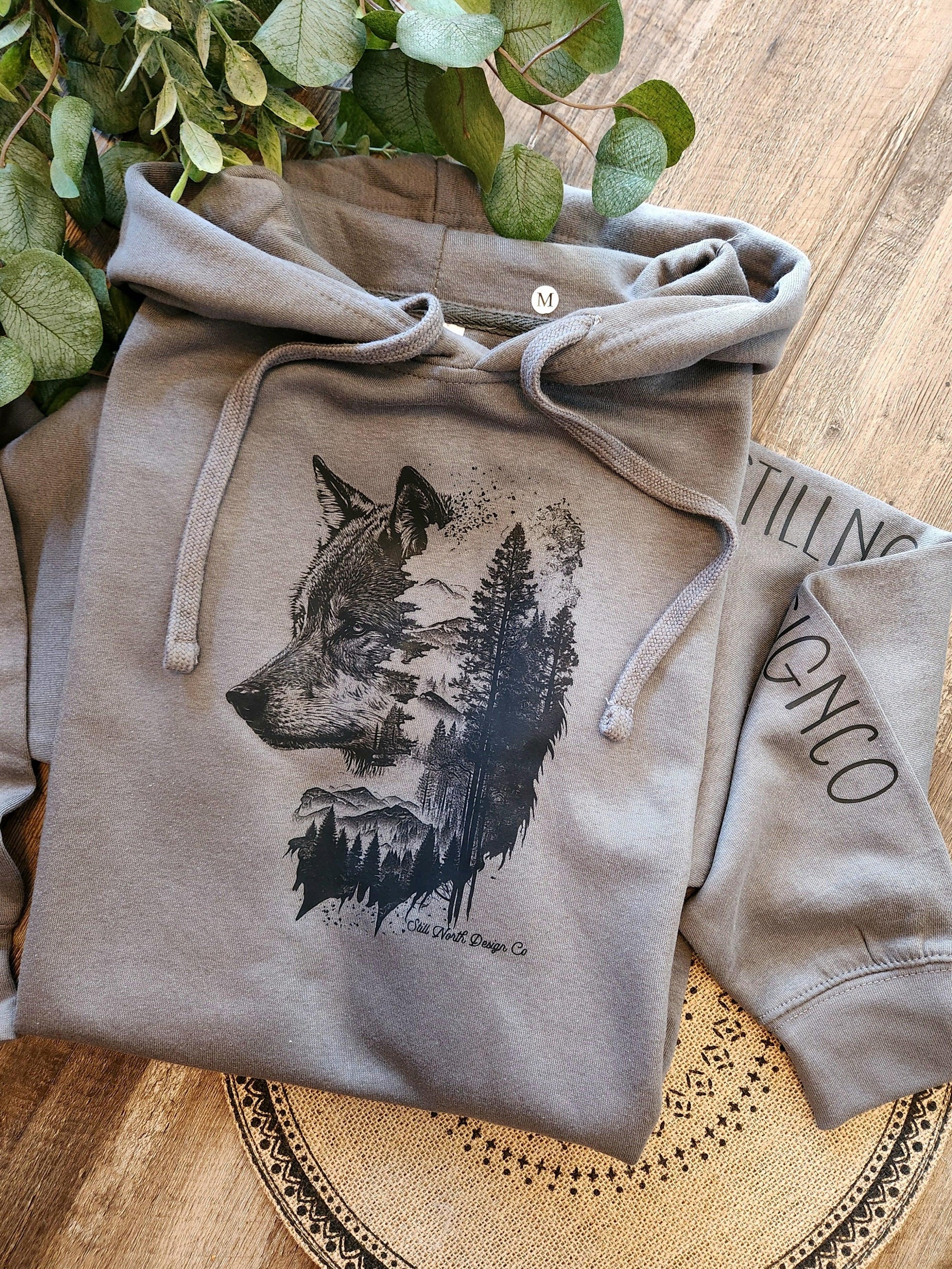 Gray hoodie with artistic wolf and forest graphic, laid on wood surface with eucalyptus leaves