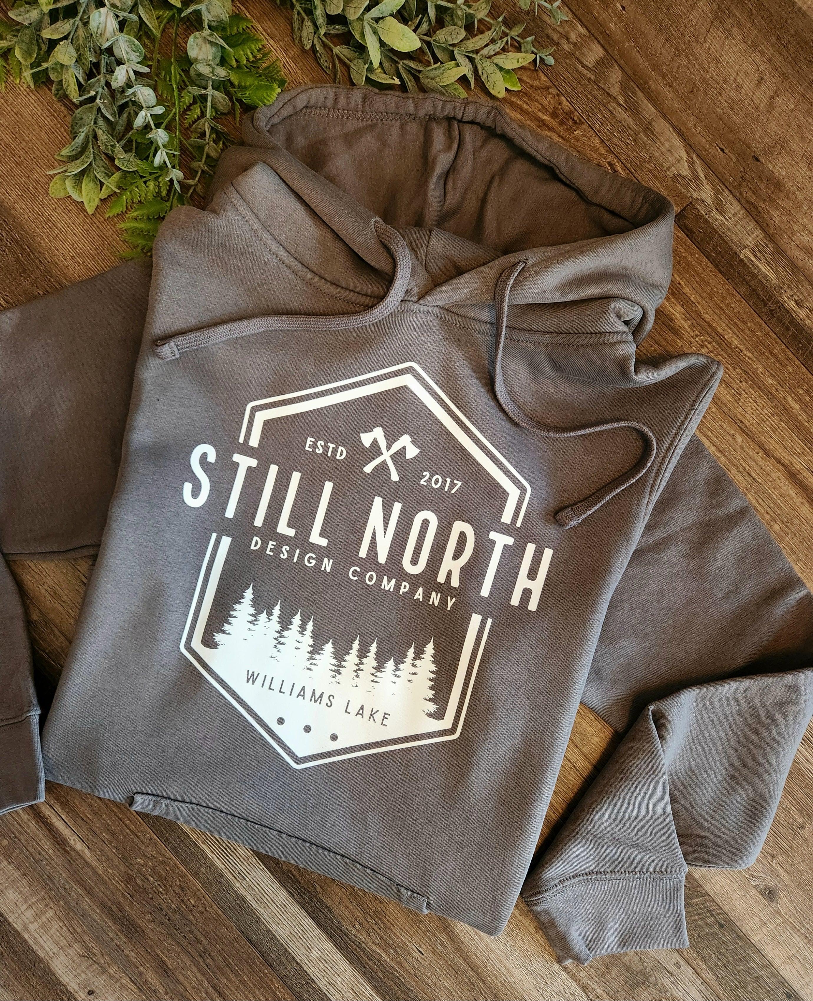 Still North Design Company hoodie on wood surface with pine tree logo and greenery accents