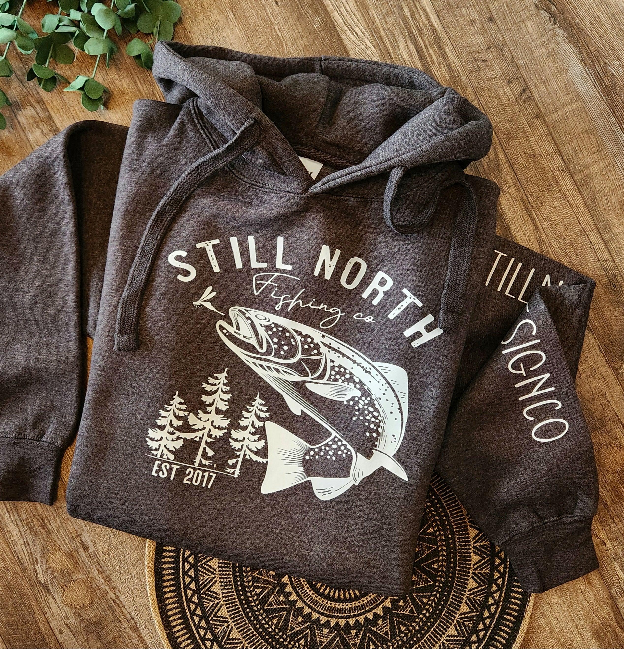 Still North Fishing Co. hoodie with fish and pine tree graphic on wood background