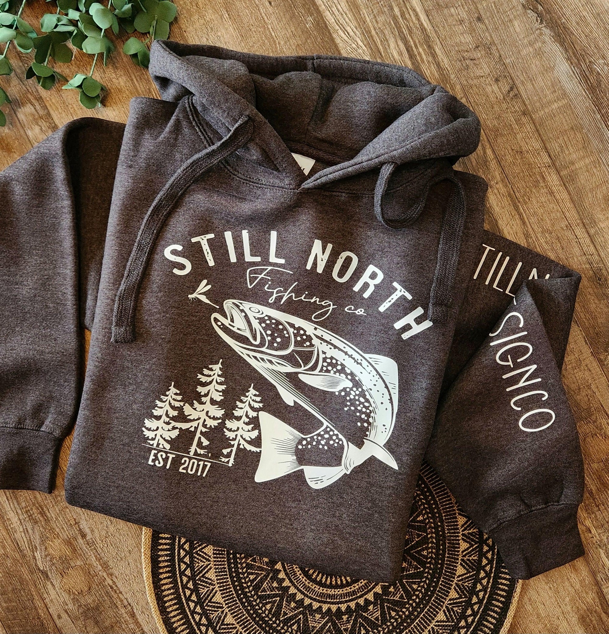 Still North Fishing Co. hoodie with fish and pine tree graphic on wood background