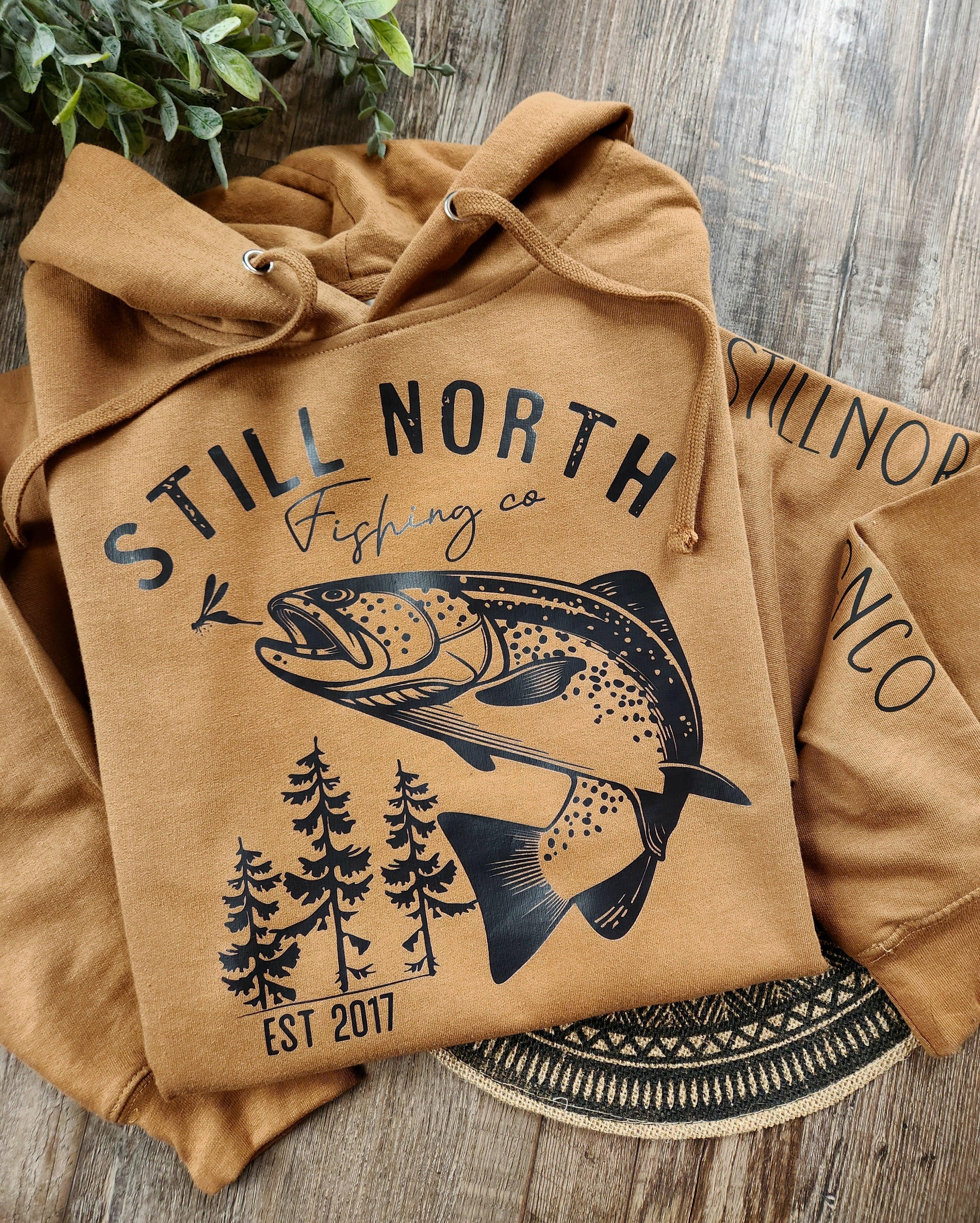 Brown Still North Fishing Co hoodie with fish and pine tree graphic on wooden surface