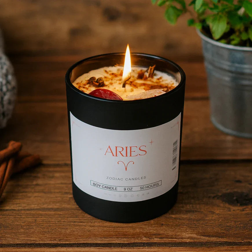 Aries zodiac soy candle burning in black jar on wooden table, plant and cinnamon beside