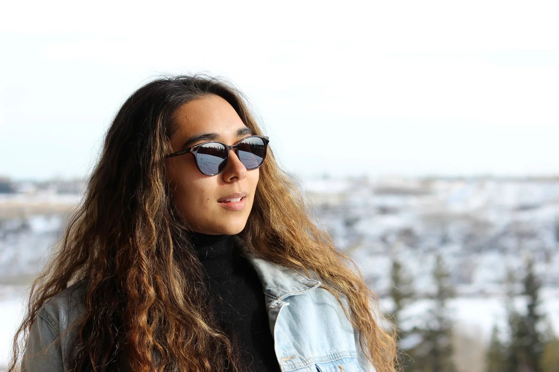 Woman with long wavy hair in sunglasses and denim jacket outdoors in snowy landscape