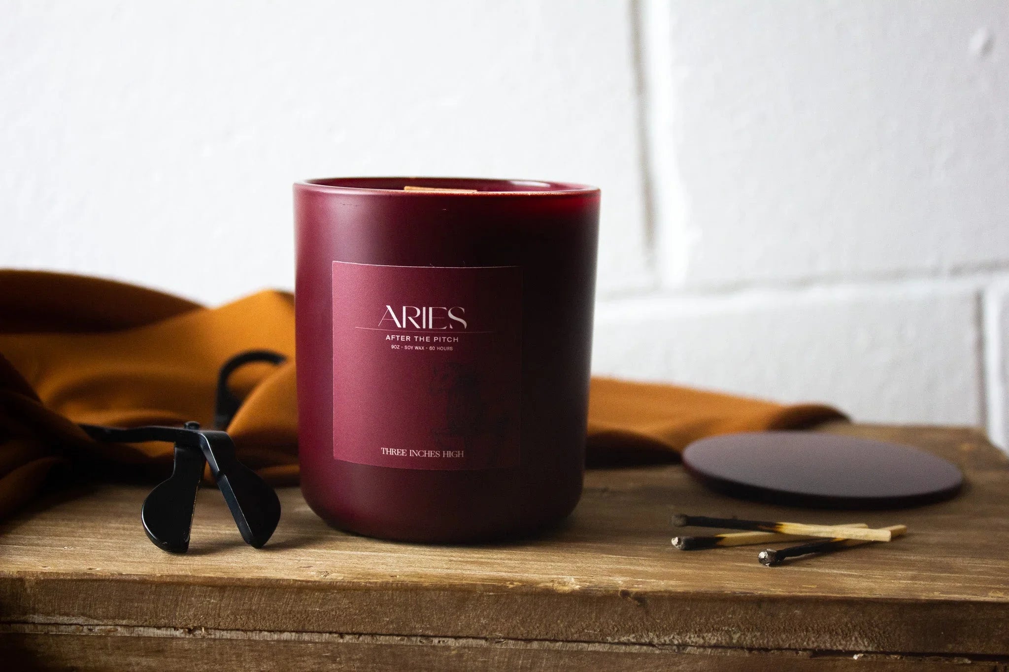 Aries soy wax candle in burgundy jar on wooden surface with matches and candle accessories