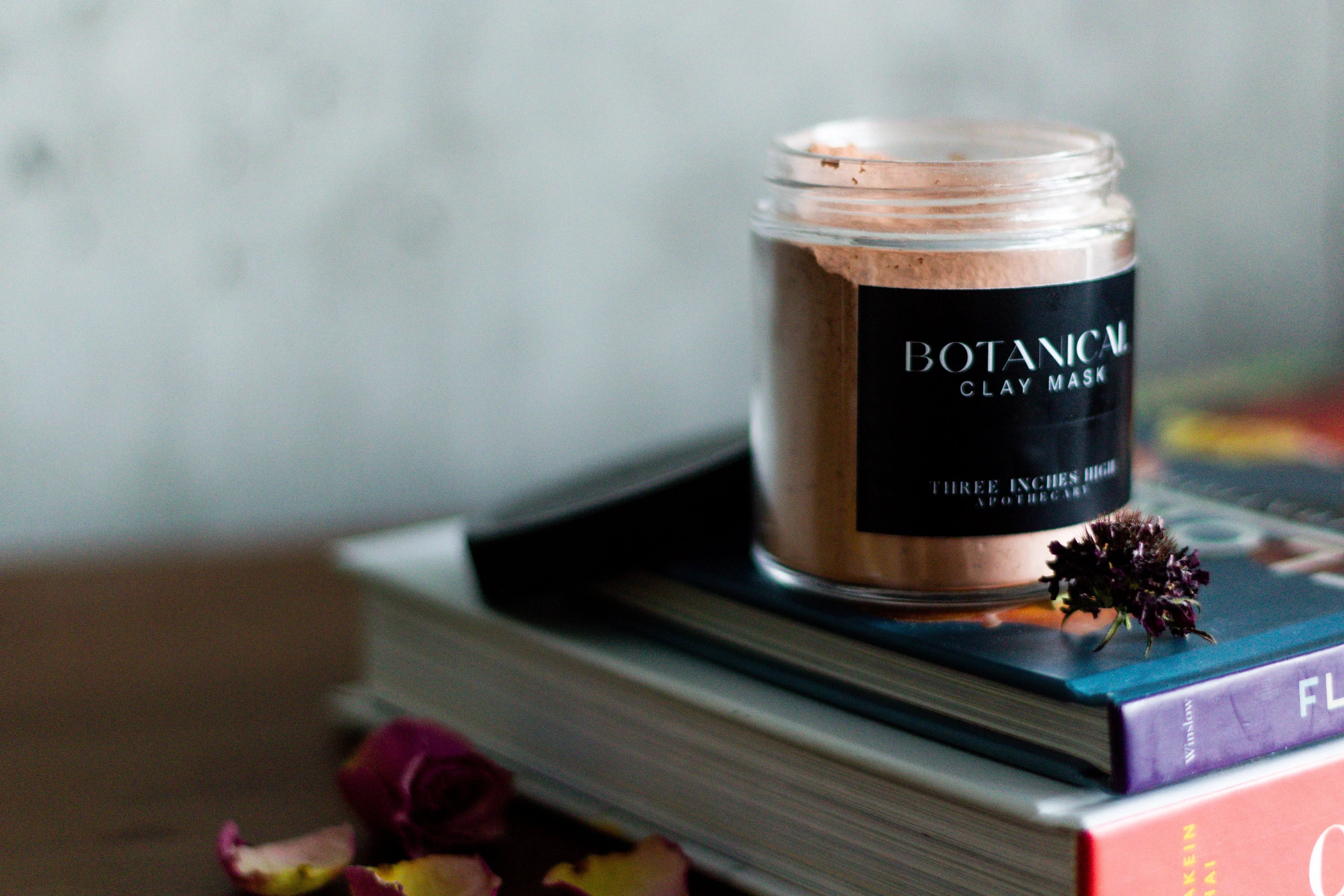 Botanical clay mask jar on stacked books with dried rose petals and flower, skincare product