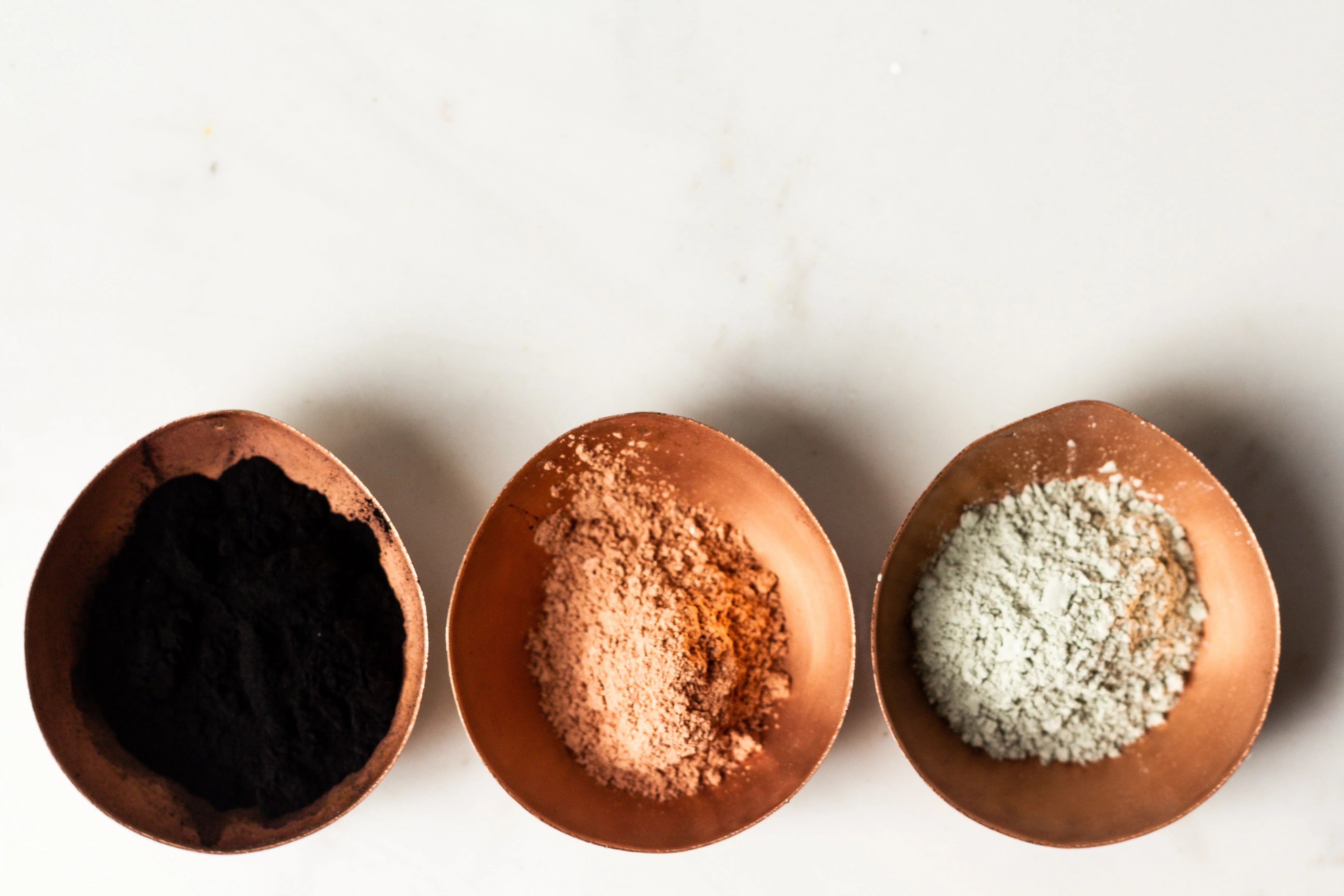 Three copper bowls with black, tan, and white powder on a white background
