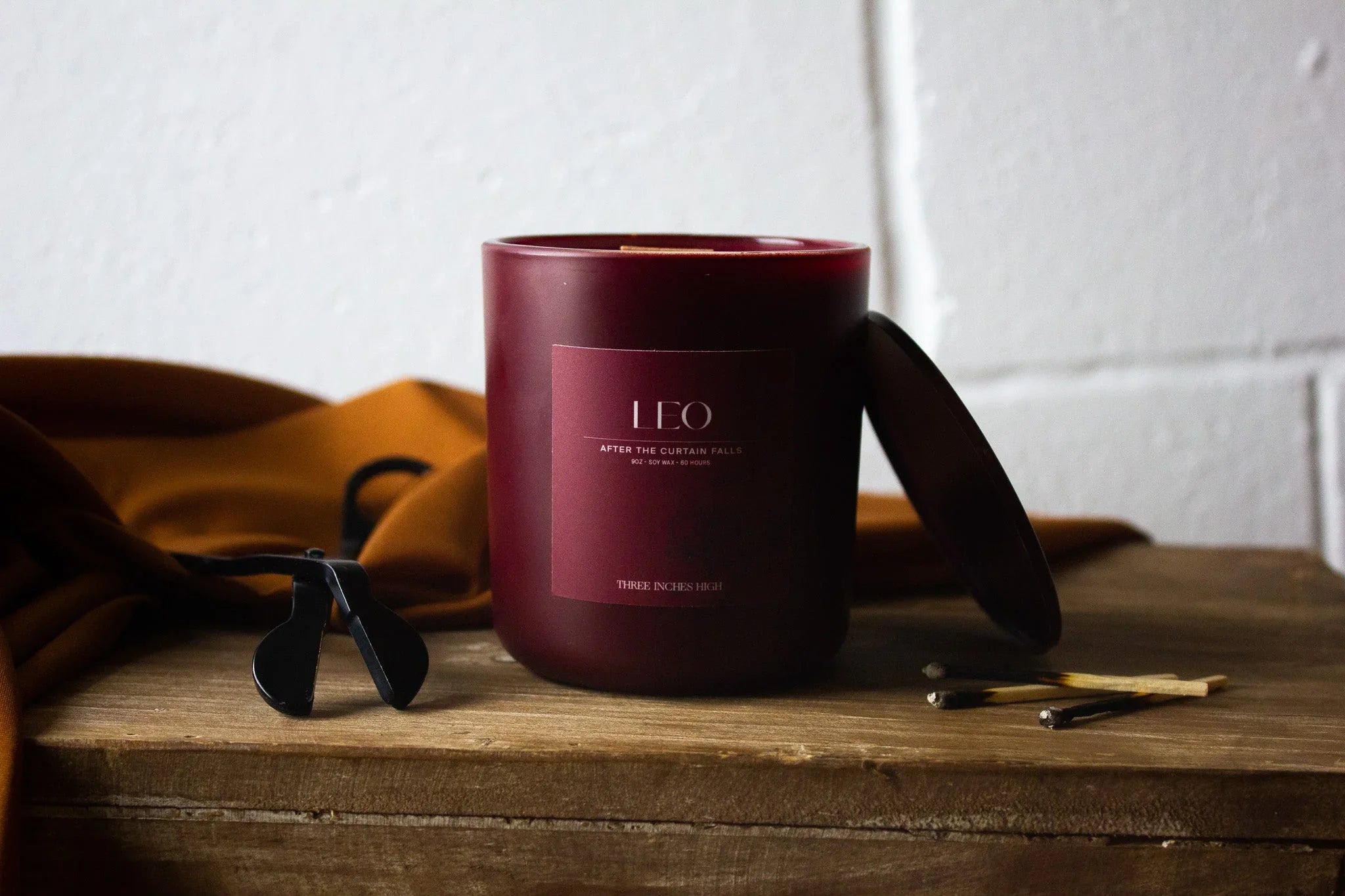 Leo scented candle in burgundy jar with lid, wick trimmer, and matches on wooden surface