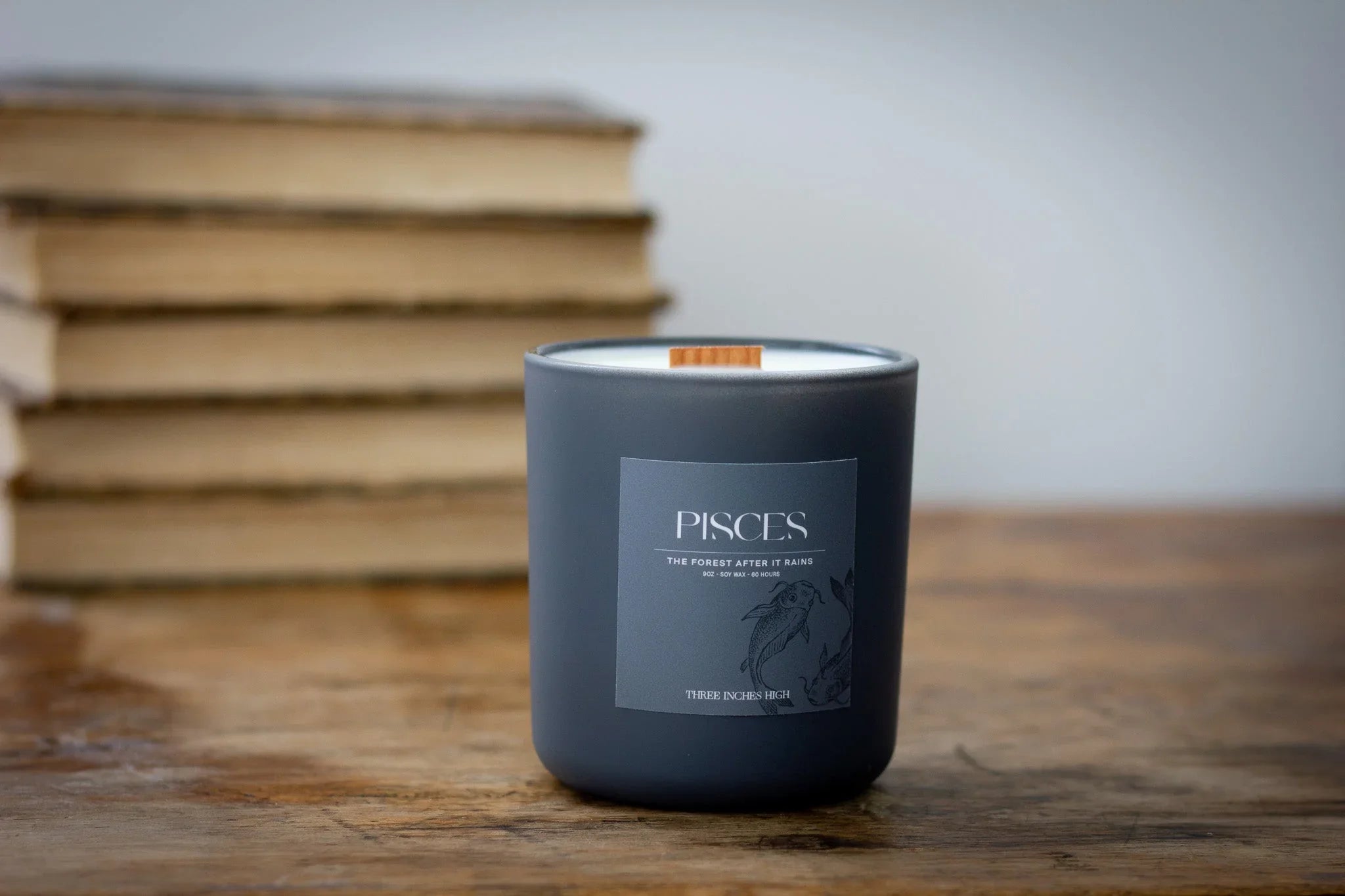 Pisces zodiac candle in matte black jar on wooden table, books stacked in background