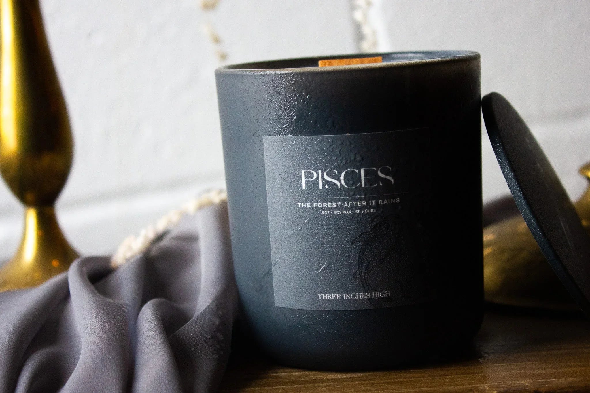 Pisces soy wax candle in matte black jar with lid on wooden table, draped fabric and brass decor