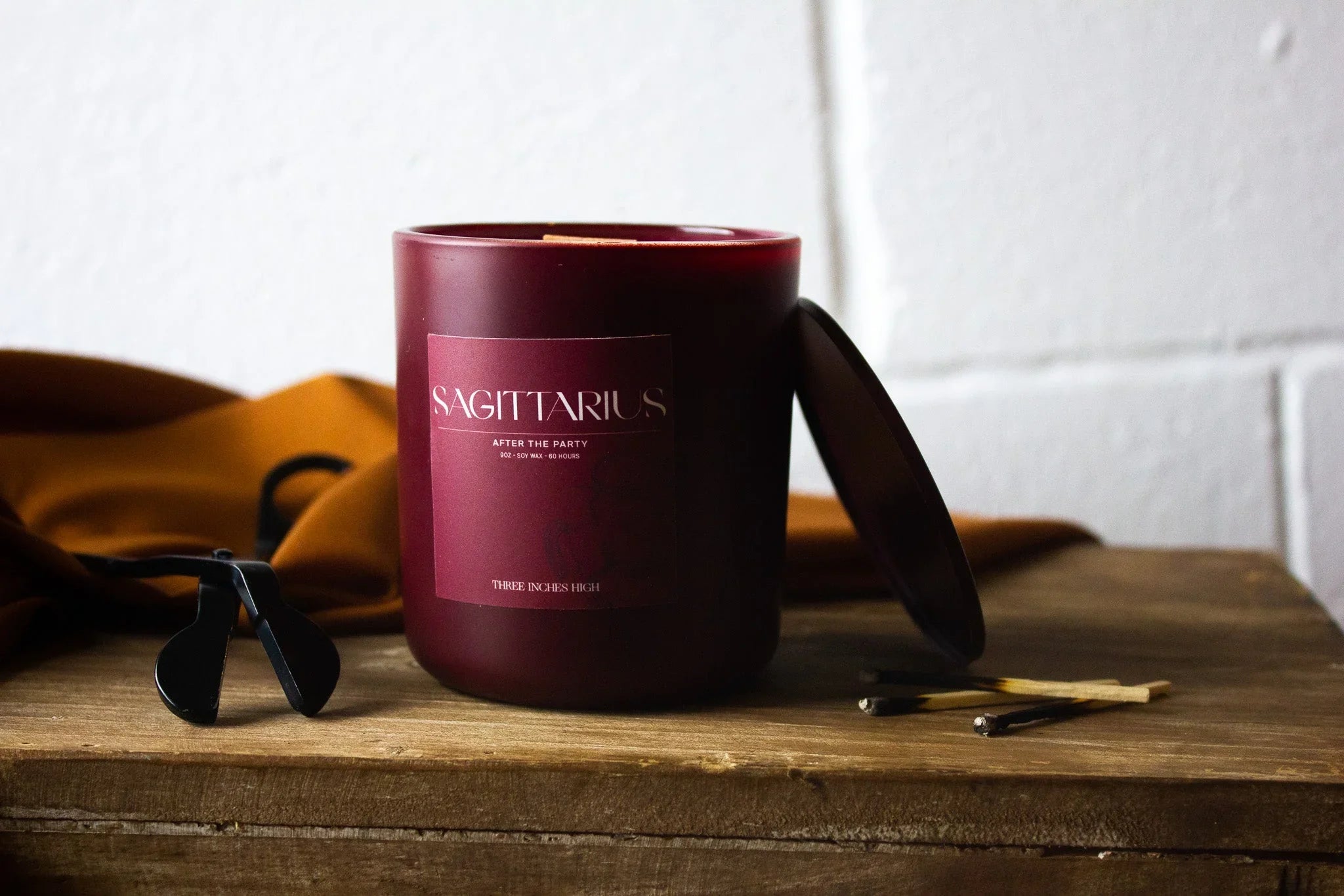 Sagittarius zodiac candle in maroon glass jar with lid, matches, and wick trimmer on wood table