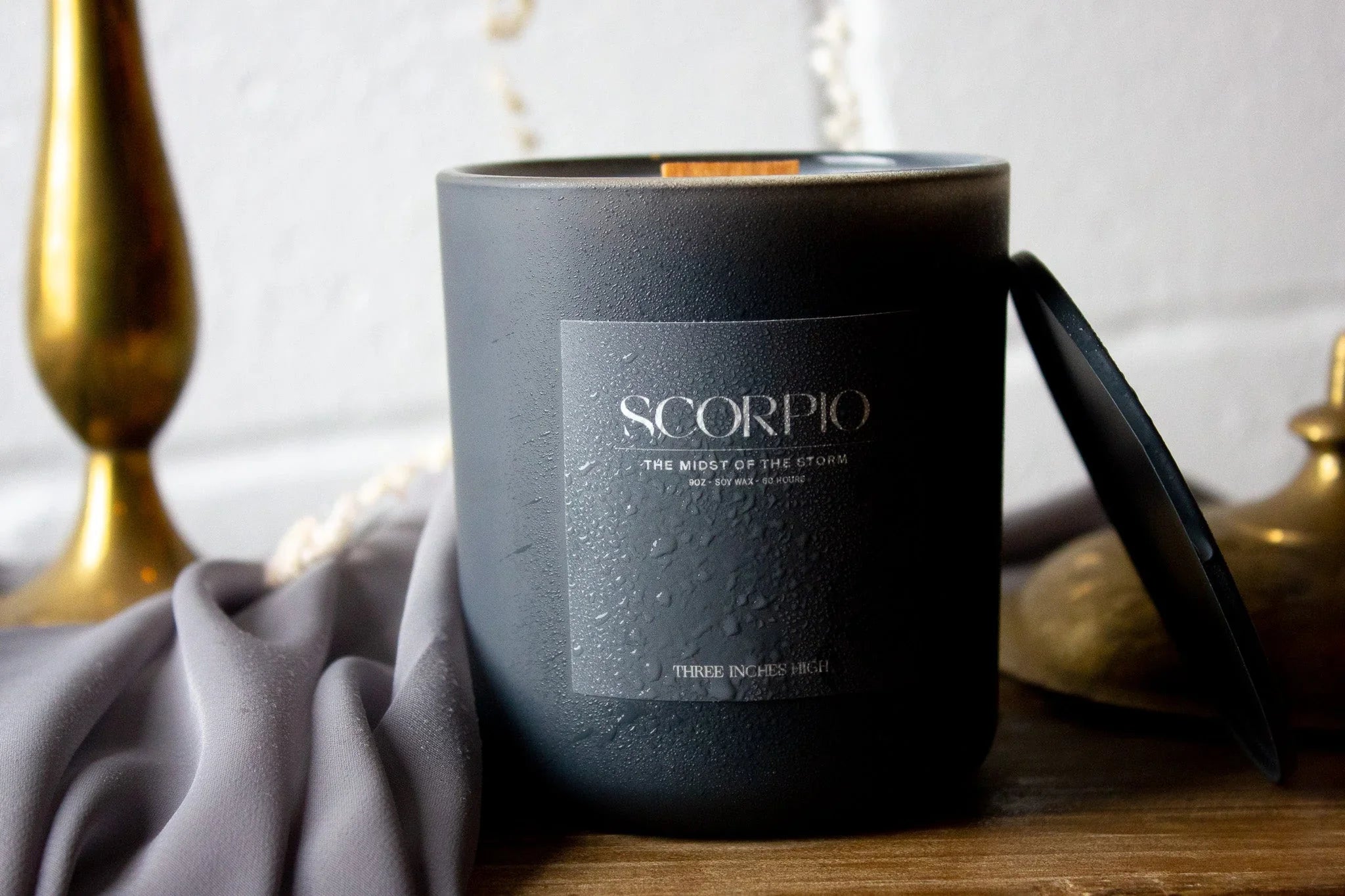 Black Scorpio soy candle with lid on wood table, gold decor and fabric backdrop.