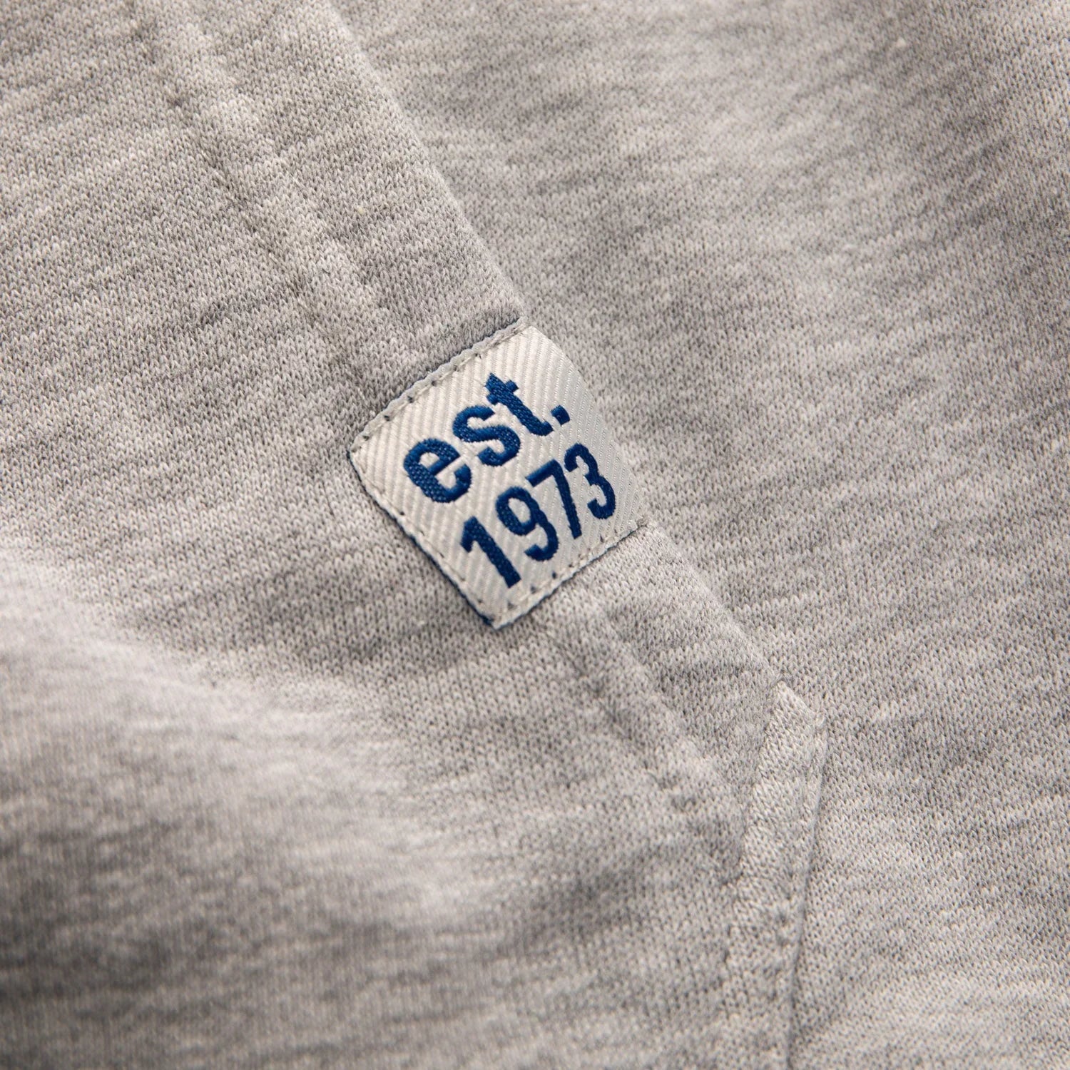 Grey sweatshirt close-up with est. 1973 blue embroidered patch near pocket